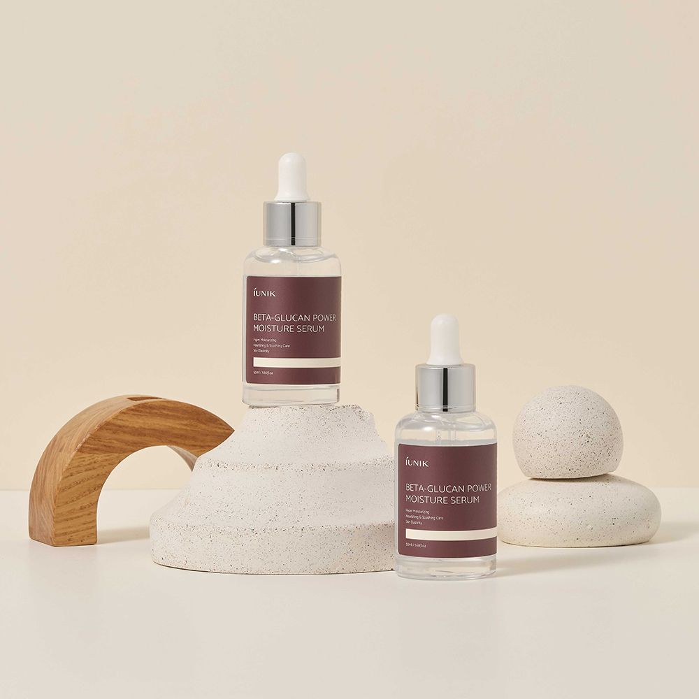 Due flaconi di siero con pipette. Scritta: IUNIK, Beta-Glucan Power Moisture Serum. Su piedistallo e accanto a pietre.