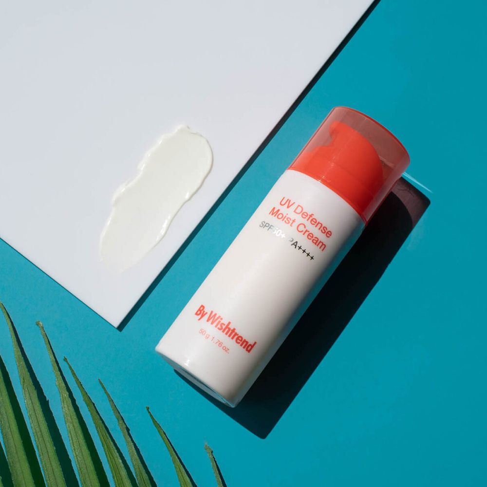 Flacone di crema solare e macchia di crema su carta bianca, sfondo blu. Marchio: By Wishtrend. Scritta: UV Defense Moist Cream SPF50+ PA++++.