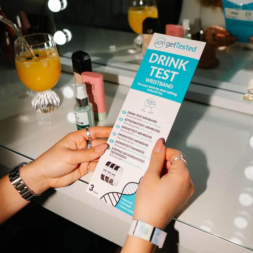 Persona che tiene una confezione con "GetTested Drink Test Wristband". Sono visibili diversi braccialetti. Sfondo: toeletta.