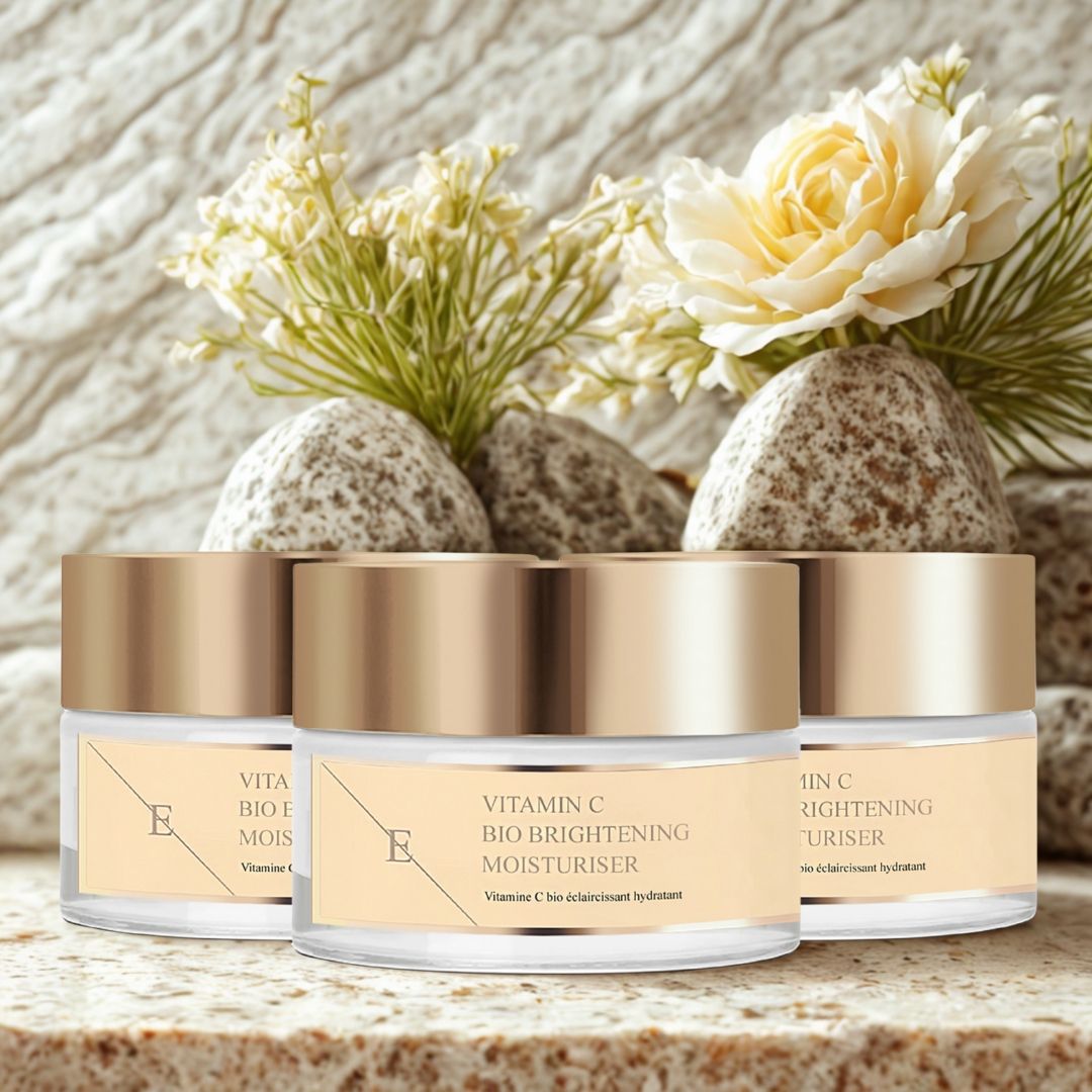 Tre vasetti con coperchi dorati, posizionati su pietre, con fiori. Le etichette riportano: Vitamin C Bio Brightening Moisturiser.