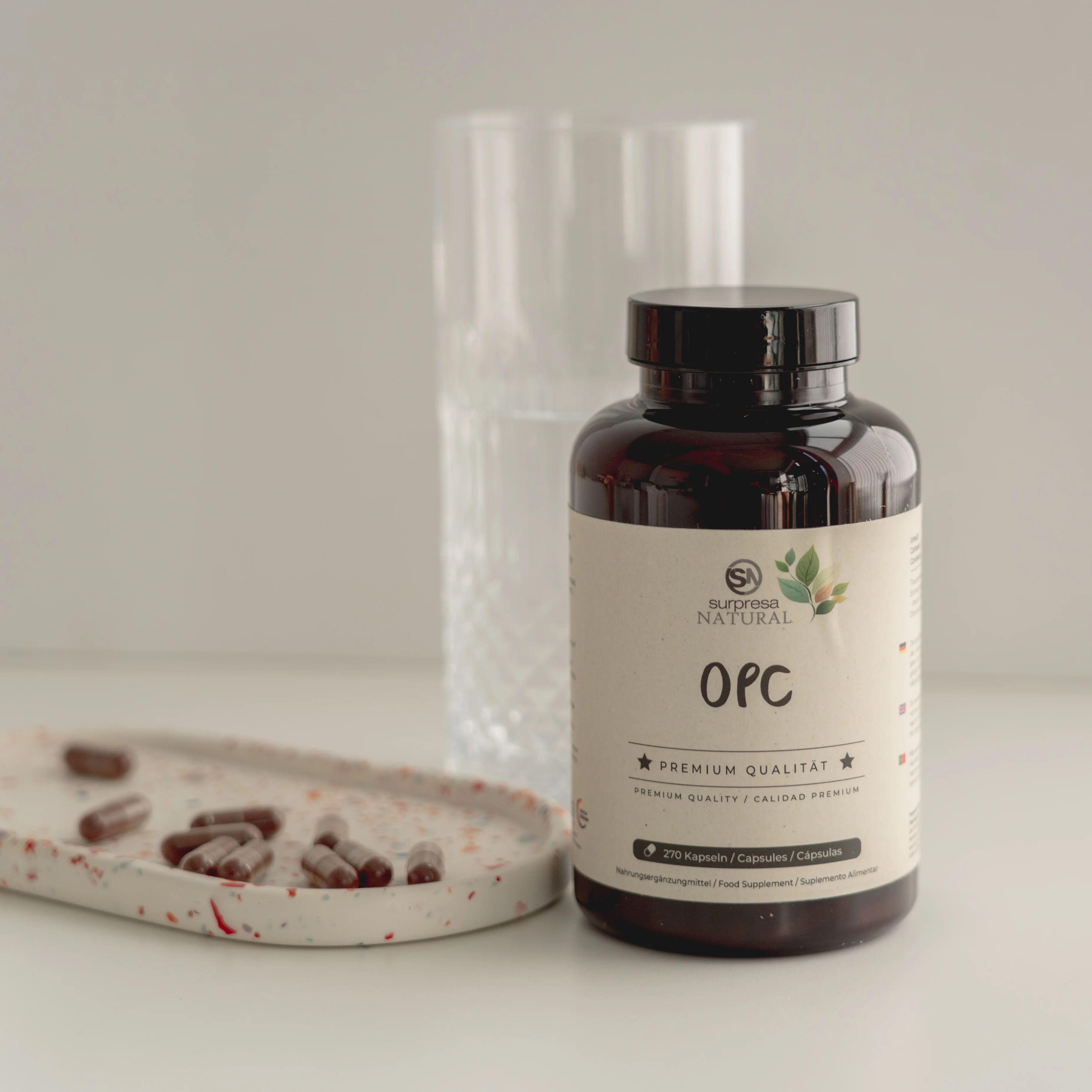Flacone marrone con etichetta 'OPC' e 'Surpresa Natural'. Accanto un bicchiere e un vassoio con capsule.