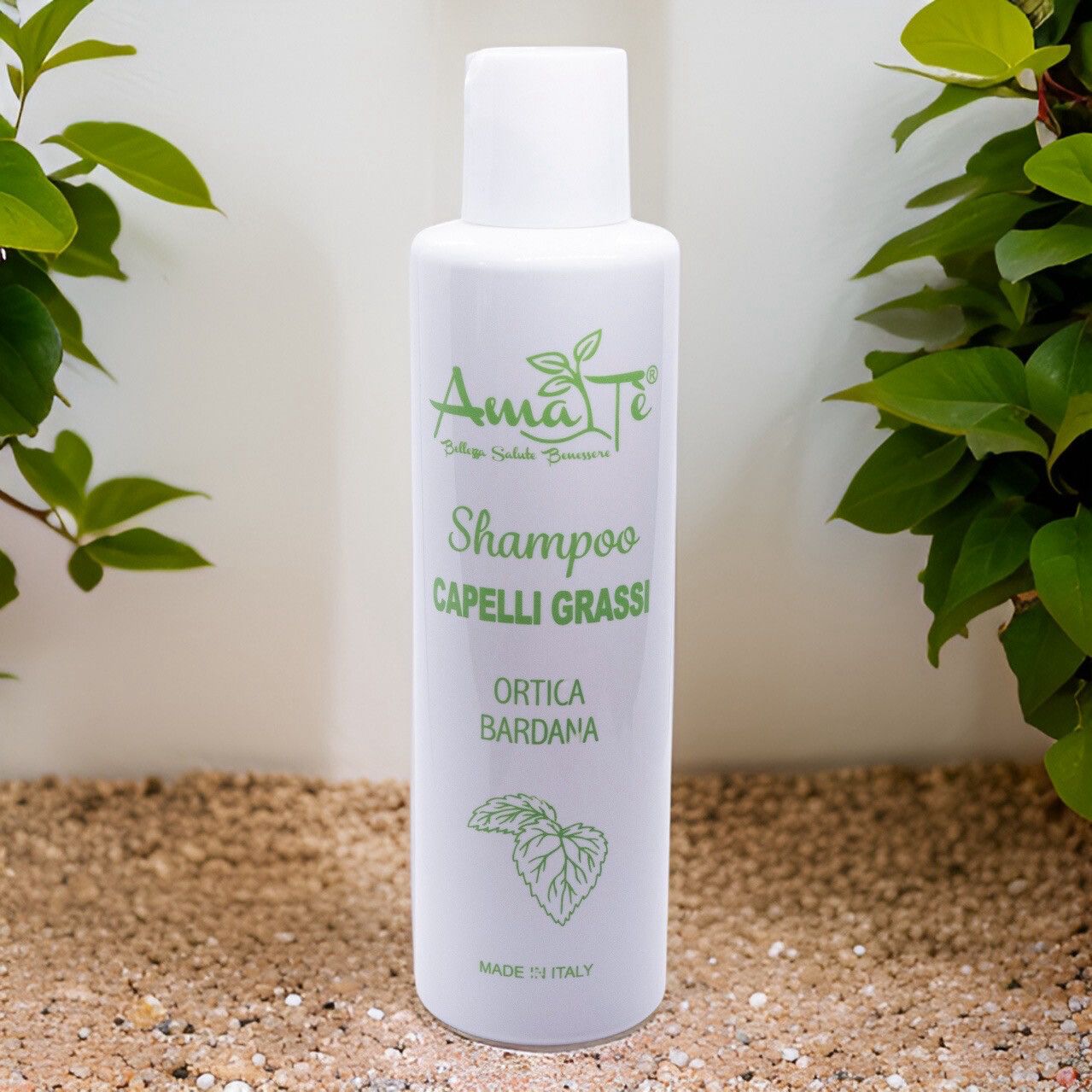 Flacone bianco con etichetta verde. Testo: Amatè Shampoo Capelli Grassi Ortica Bardana. Sfondo: sabbia.