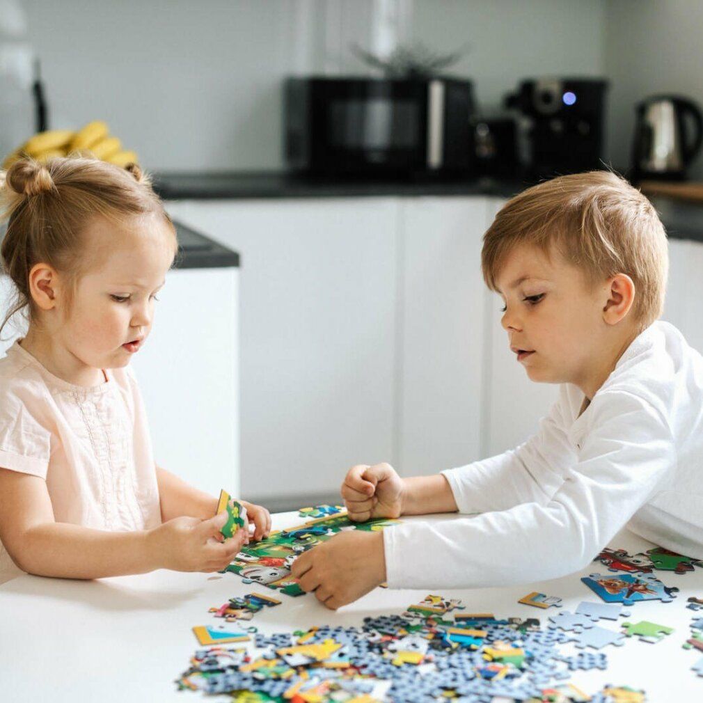 Due bambini seduti a un tavolo compongono un puzzle. Sul tavolo ci sono pezzi di puzzle. Entrambi i bambini indossano abiti chiari.