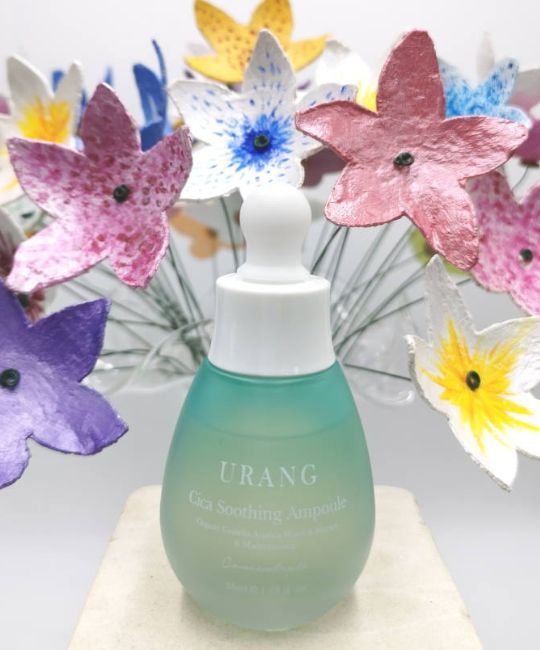 Flacone contagocce, tappo bianco. Vetro turchese opaco. Scritta: URANG Cica Soothing Ampoule. Davanti a fiori.