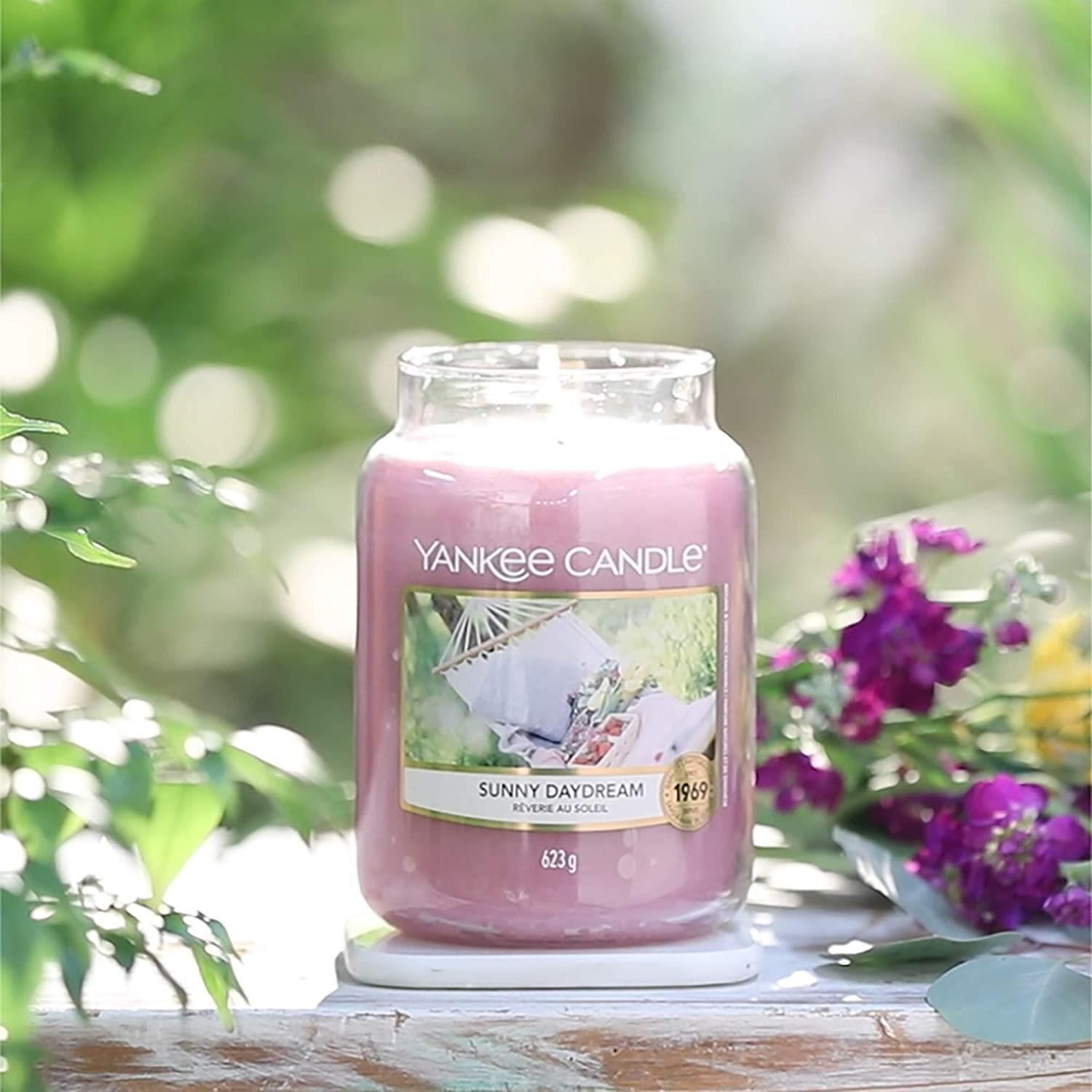 Candela profumata Yankee Candle Sunny Daydream. Candela rosa in barattolo di vetro. Etichetta con immagine amaca. Peso: 623 g. Su sottobicchiere bianco.
