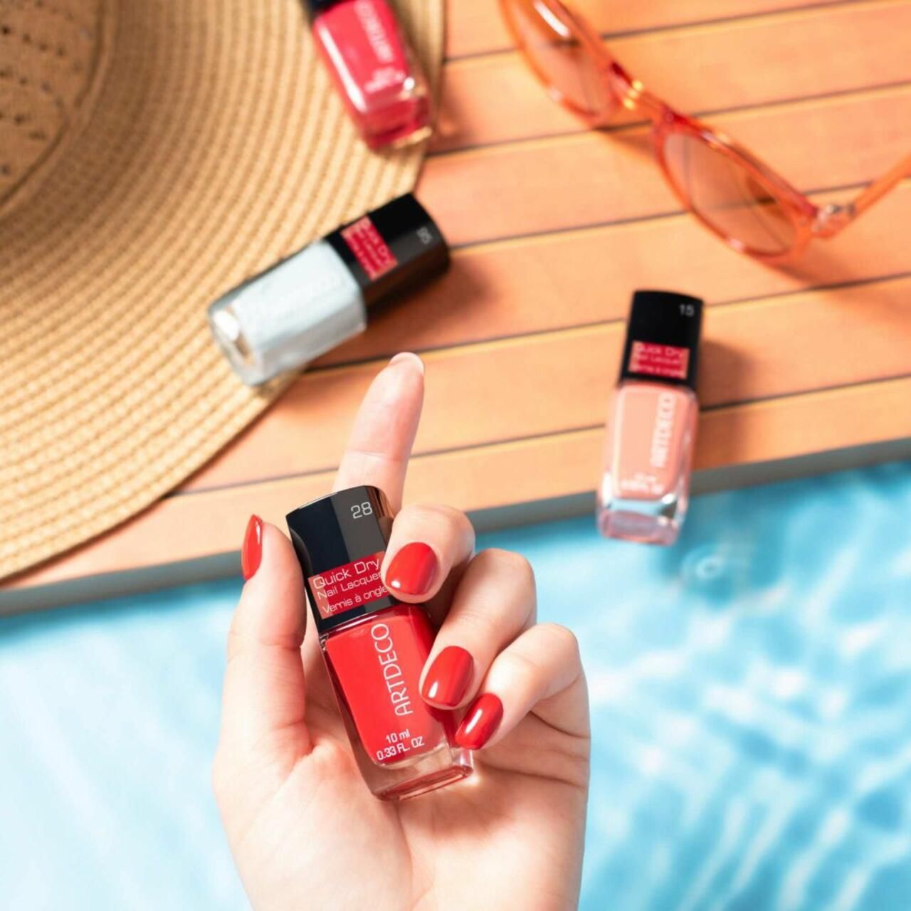 Mano che tiene un flacone di smalto per unghie Artdeco Quick Dry rosso. Altri flaconi e occhiali da sole. Unghie smaltate.