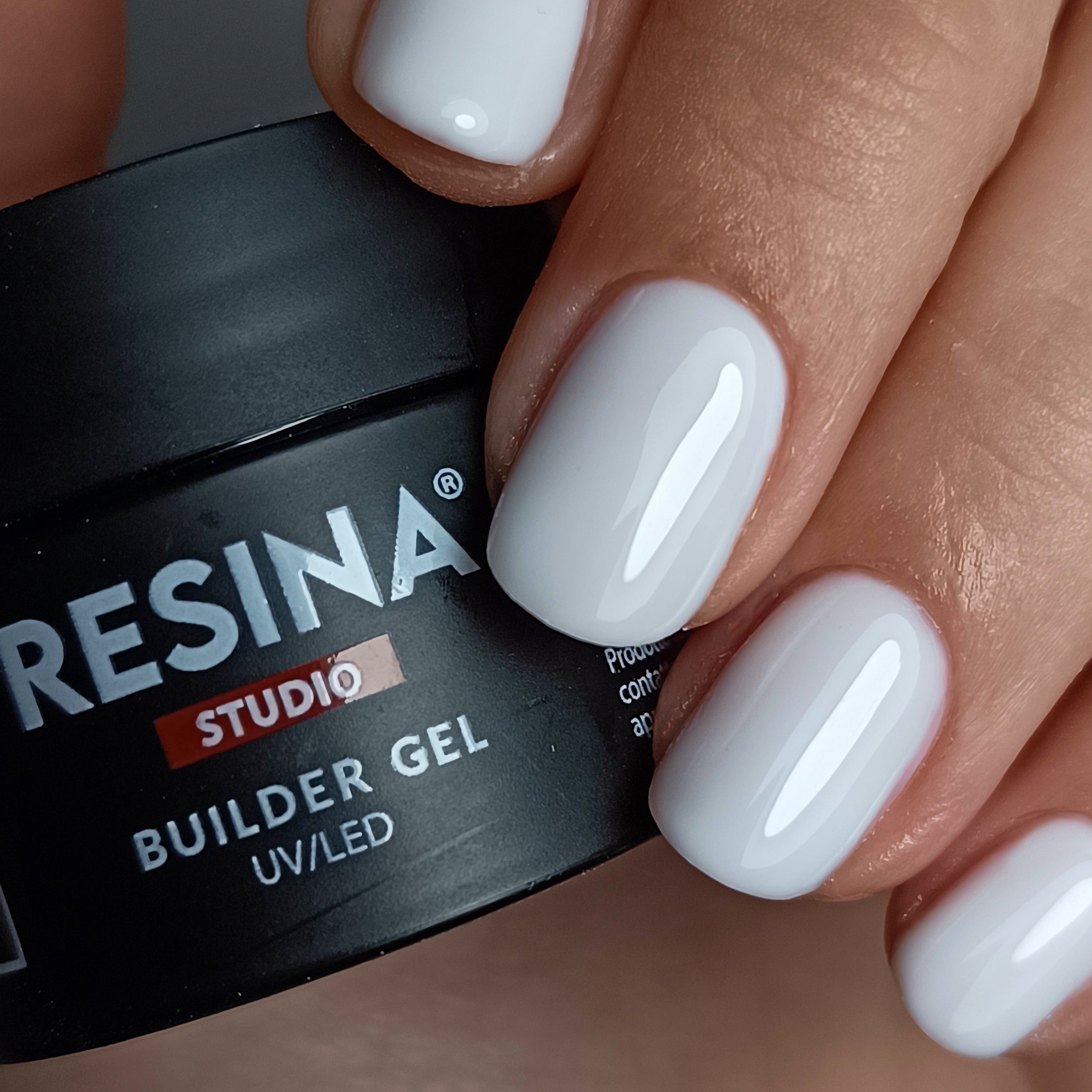 Mano con unghie smaltate. Scritta: RESINA STUDIO, BUILDER GEL, UV/LED. Unghie bianco latte.