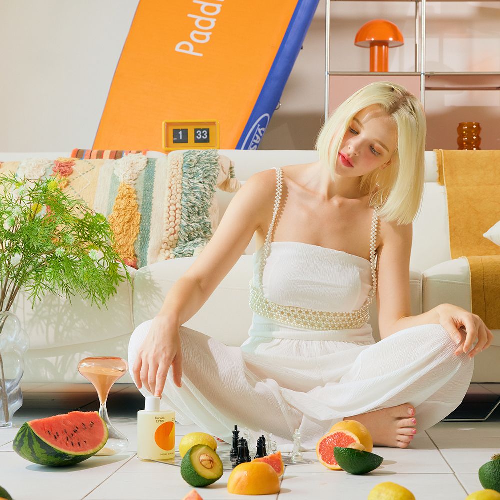 Una donna seduta a terra con frutta e un flacone di bagnoschiuma. Sul flacone sono visibili testo e un disegno arancione.