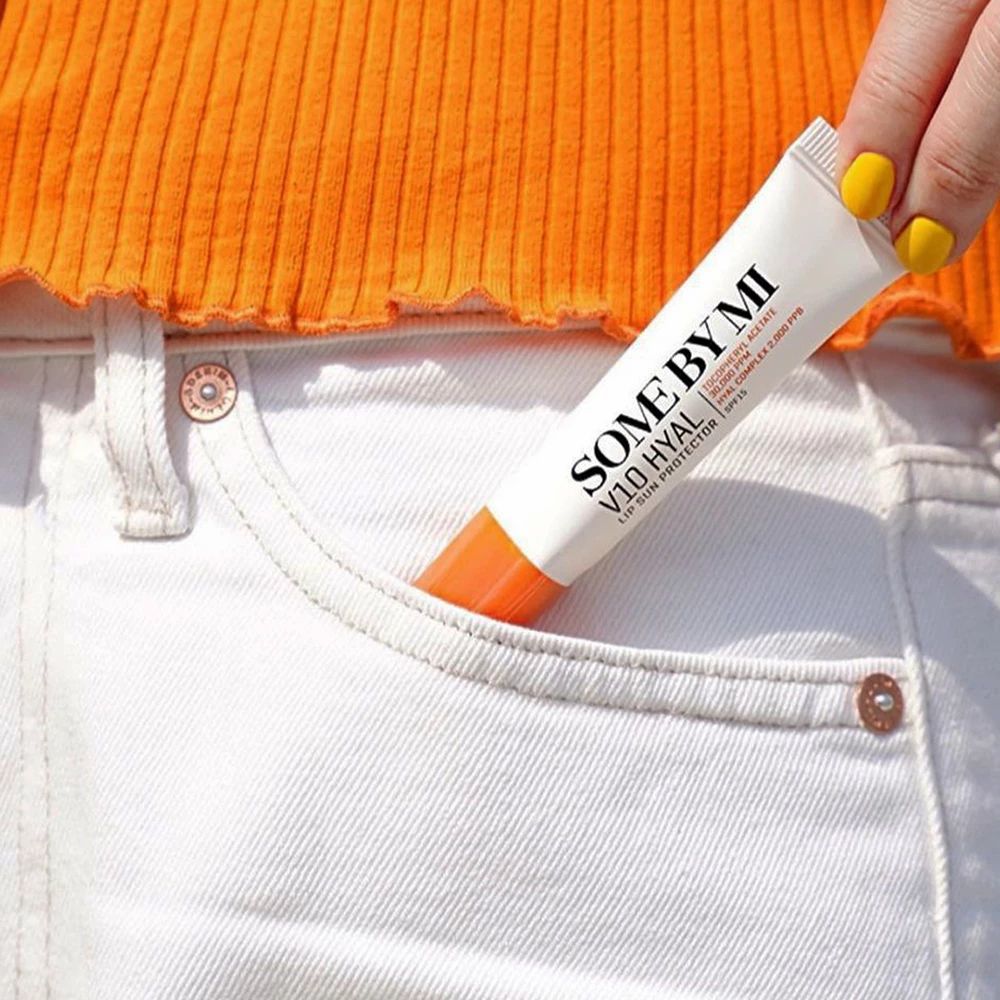 Tubo SOME BY MI V10 Hyal Lip Protector in tasca. Persona con top arancione e jeans bianchi. Unghie gialle.