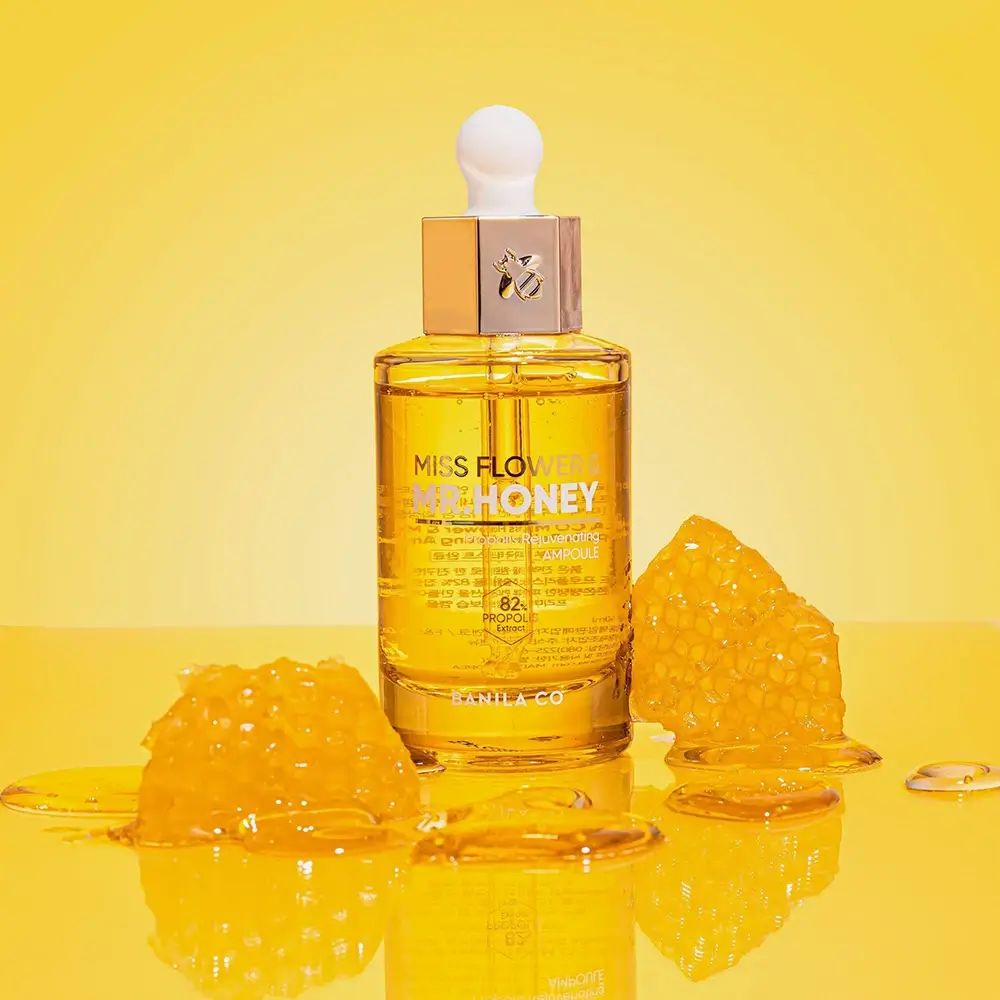 Bottiglia di vetro con siero dorato. Scritta: Miss Flower & Mr. Honey. Favo accanto. Sfondo giallo.
