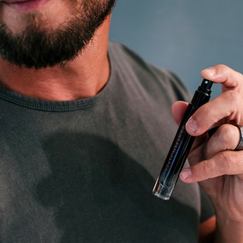 Primo piano di un uomo che spruzza profumo. Flacone nero con scritta rossa. L'uomo indossa una maglietta grigia.