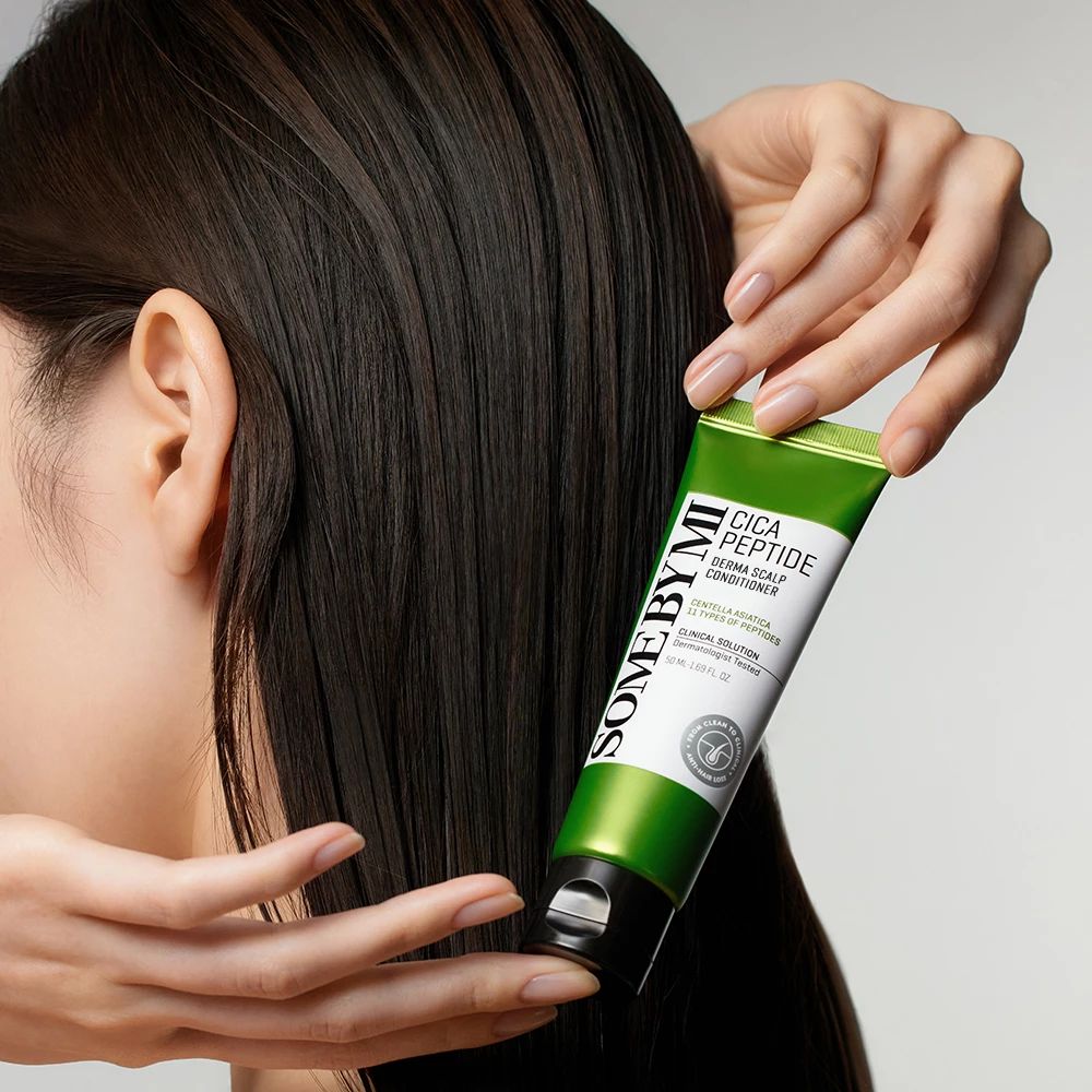 Tubo verde SOME BY MI Cica Peptide Derma Scalp Conditioner. Mani che tengono il tubo vicino a capelli scuri.