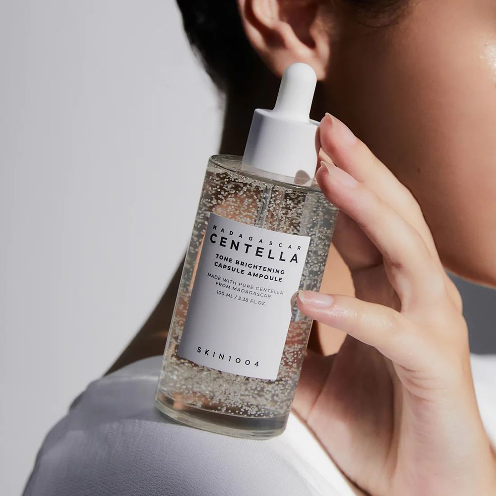 Persona che tiene in mano flacone di siero con pipetta. Flacone con etichetta bianca. Marchio: SKIN1004. Nome prodotto: Madagascar Centella Tone Brightening Ampoule.