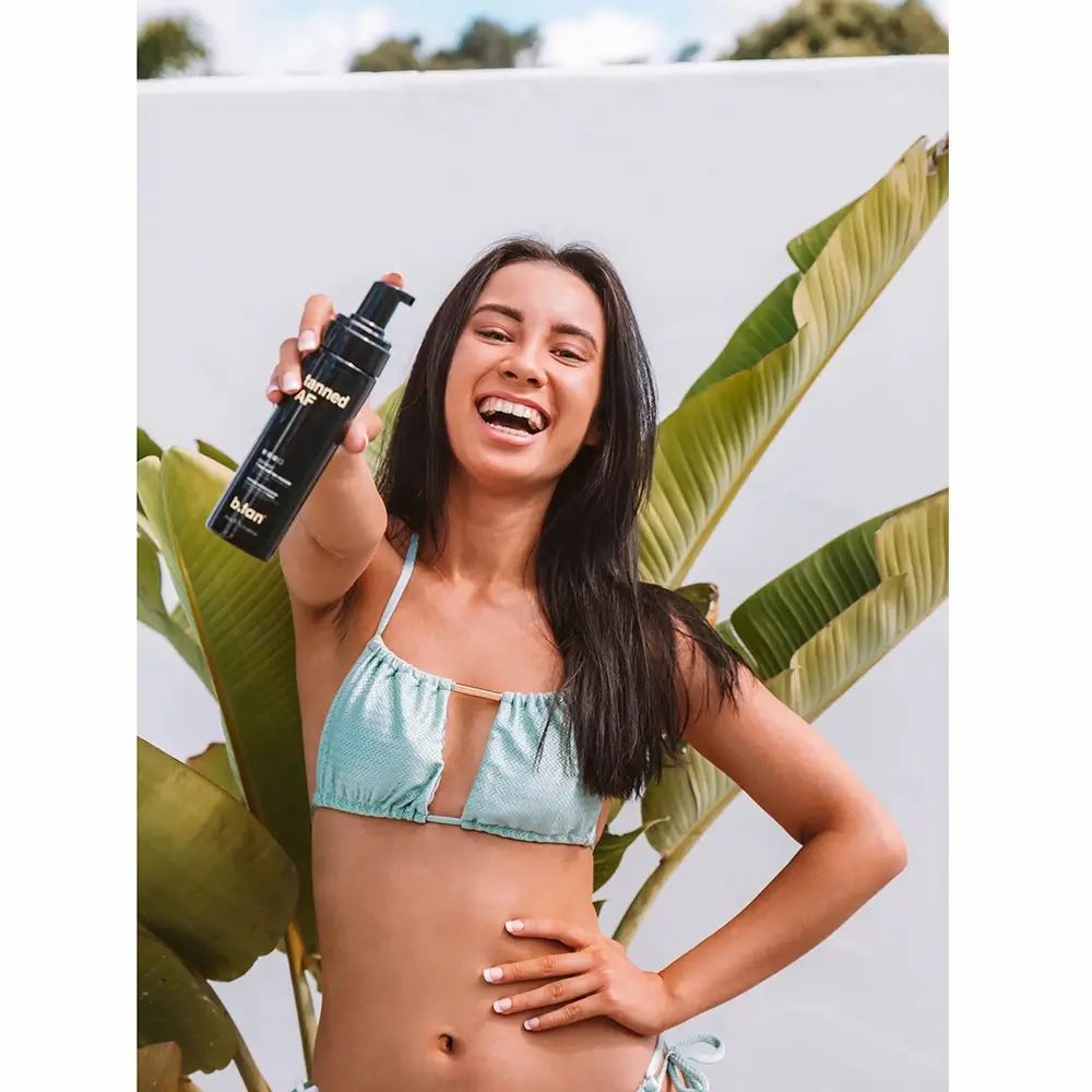 Una donna tiene in mano un flacone nero con testo giallo. Scritta: Tanned AF. Indossa un bikini e si trova davanti a un muro e delle foglie.