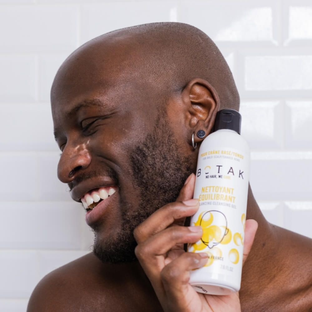 Uomo tiene una bottiglia. Scritta BOTAK, NETTOYANT ÉQUILIBRANT. Sorride. Orecchino nero.