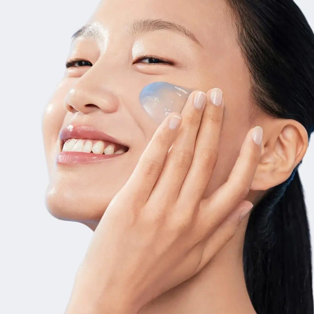 Donna applica crema blu sul viso. Sorriso. Pelle chiara. Capelli neri. Sfondo azzurro.