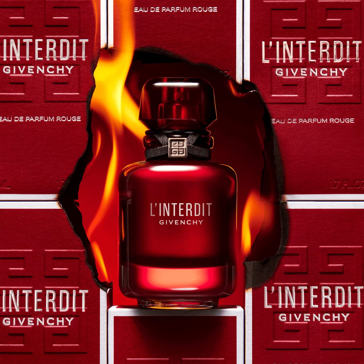 Flacone di profumo rosso con nastro nero e tappo argentato, circondato da fiamme e scatole rosse con scritta.
