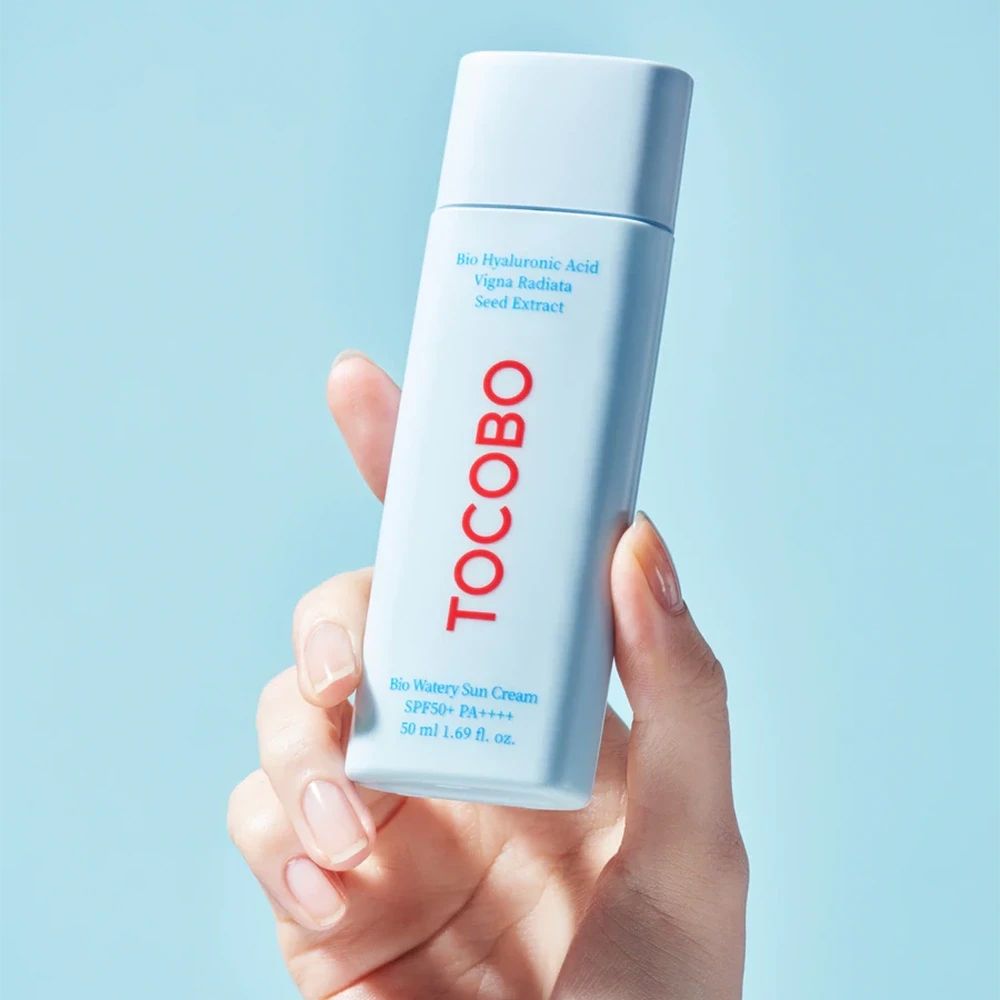 Mano che tiene un tubo azzurro. Scritta TOCOBO rossa. Testo: Bio Watery Sun Cream SPF50+ PA++++. 50 ml.