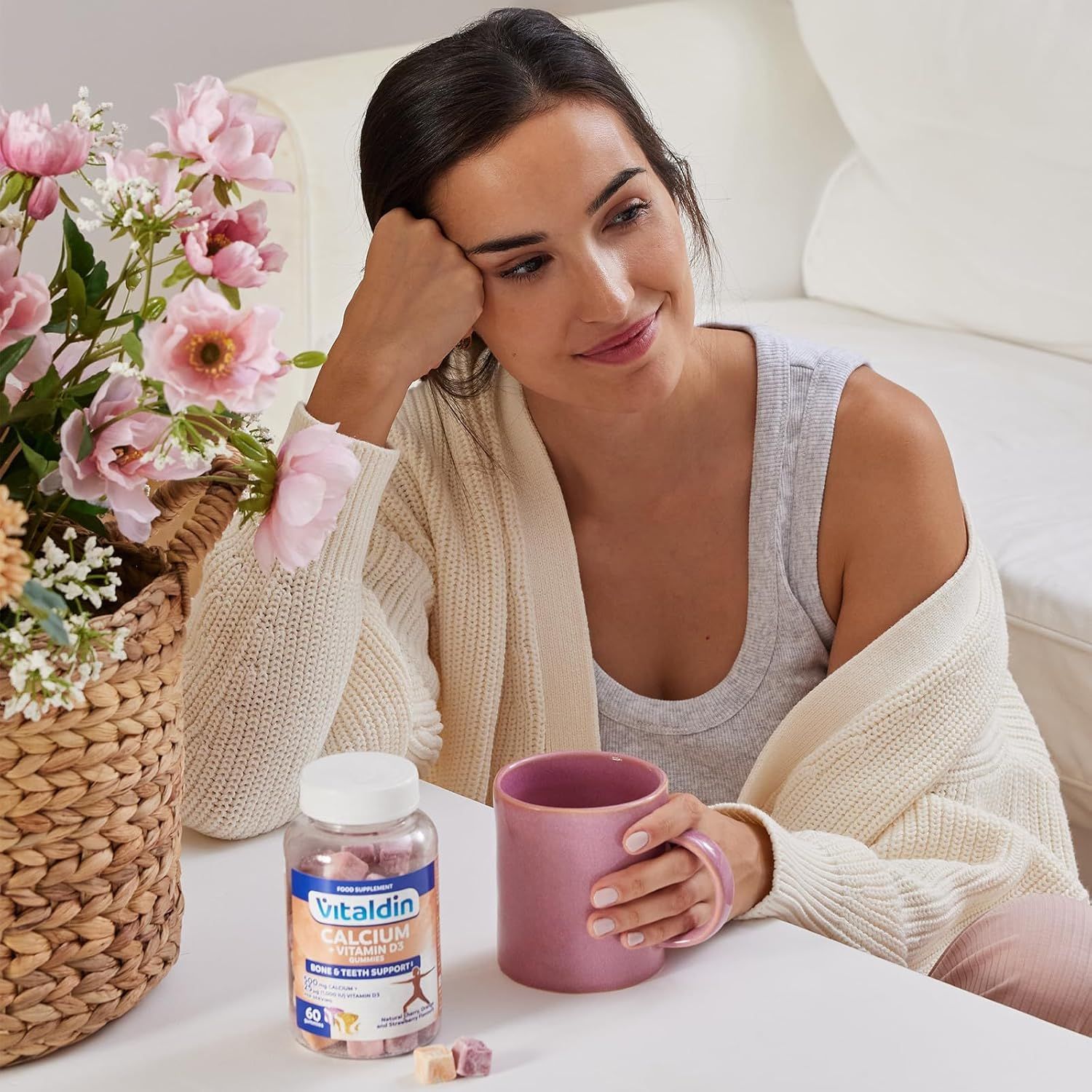 Donna seduta con flacone VITALDIN Calcium + Vitamina D3 Gummies e tazza. 60 caramelle gommose. Caramelle gommose sul tavolo. Tazza rosa.