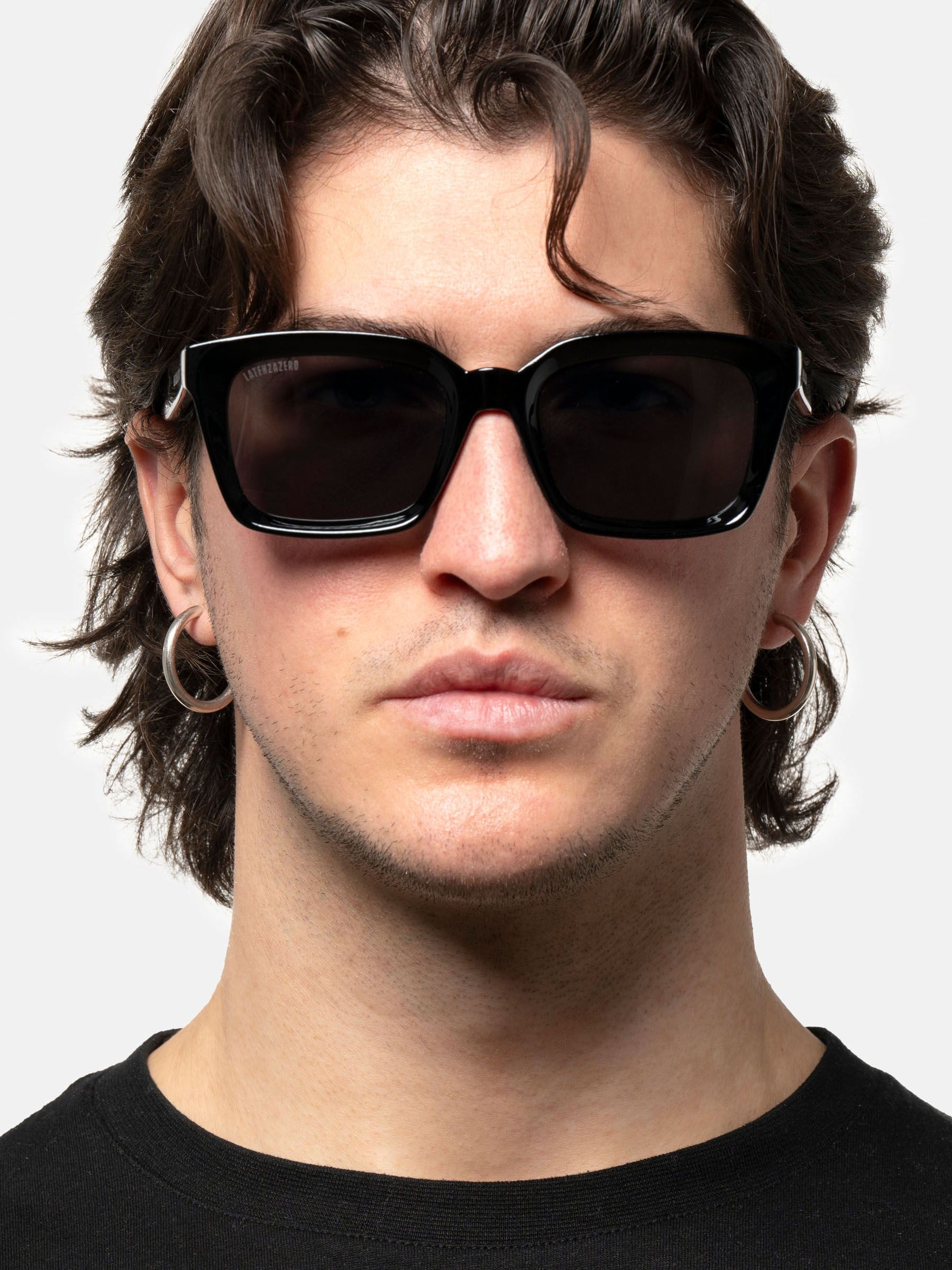 Uomo con occhiali da sole neri. Lenti quadrate, montatura nera. Marchio sulla montatura. Capelli corti.