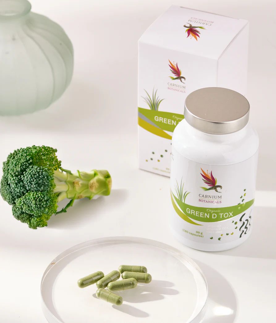Flacone e scatola bianchi con logo e "GREEN D TOX". Capsule verdi su piatto di vetro. Broccoli.