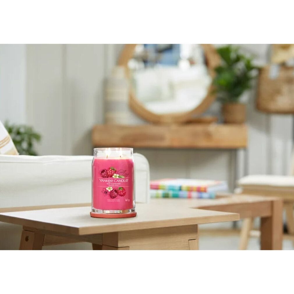 Candela profumata rossa su un tavolo. Sullo sfondo: divano, specchio e decorazioni. Scritta: Red Raspberry.