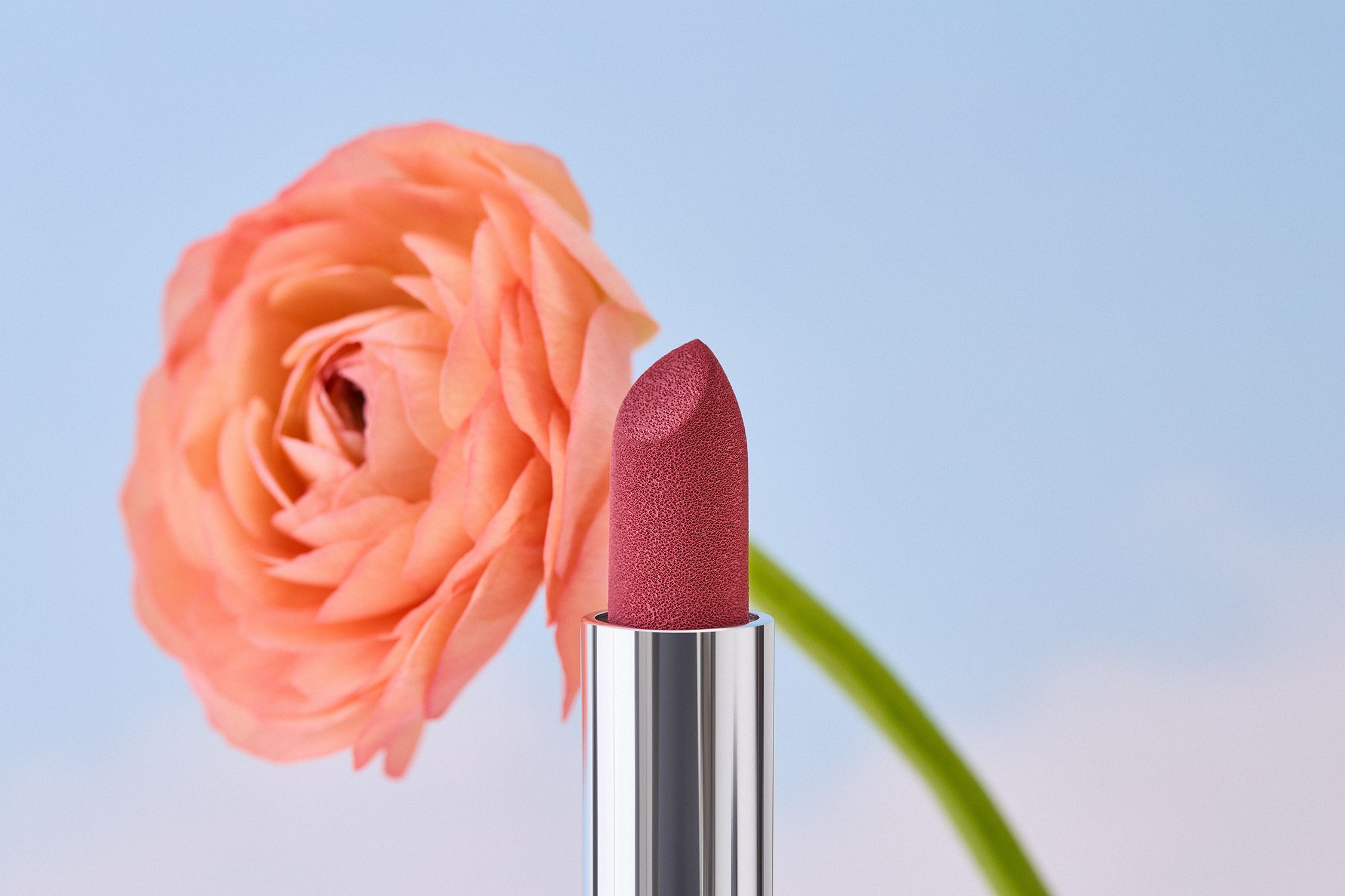 Rossetto rosa davanti a un fiore arancione. Astuccio argentato, punta del rossetto visibile.