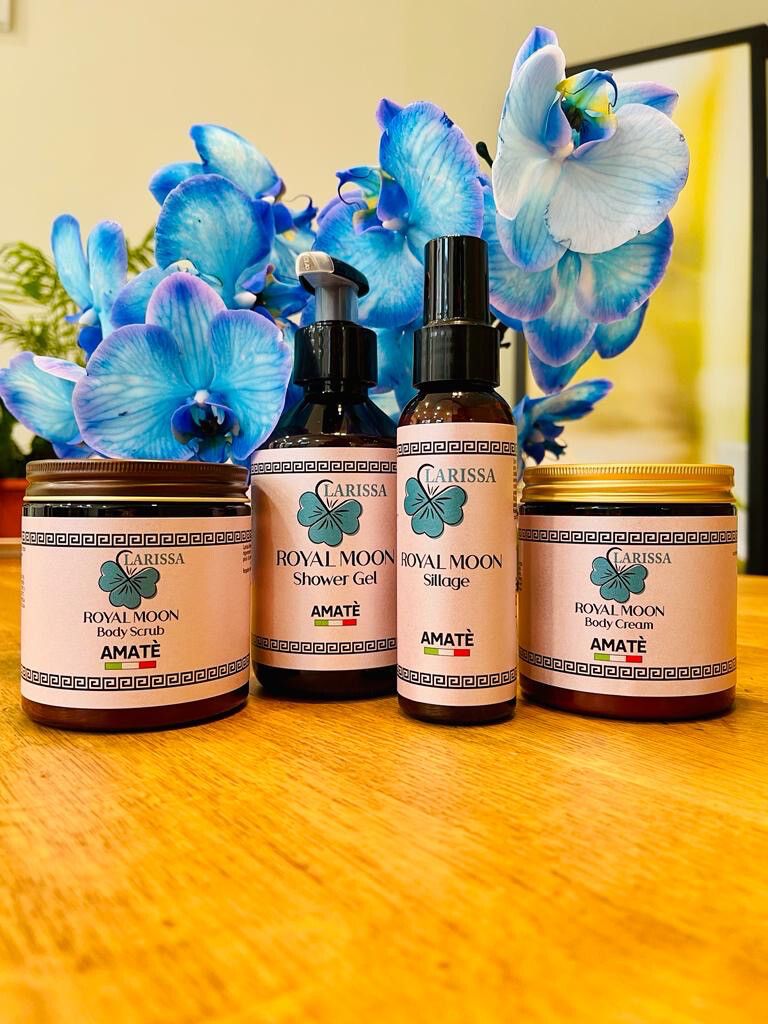 Quattro prodotti con etichette "Larissa Royal Moon" e "Amatè". Contiene: Body Scrub, Shower Gel, Sillage e Body Cream. Davanti a orchidee blu.