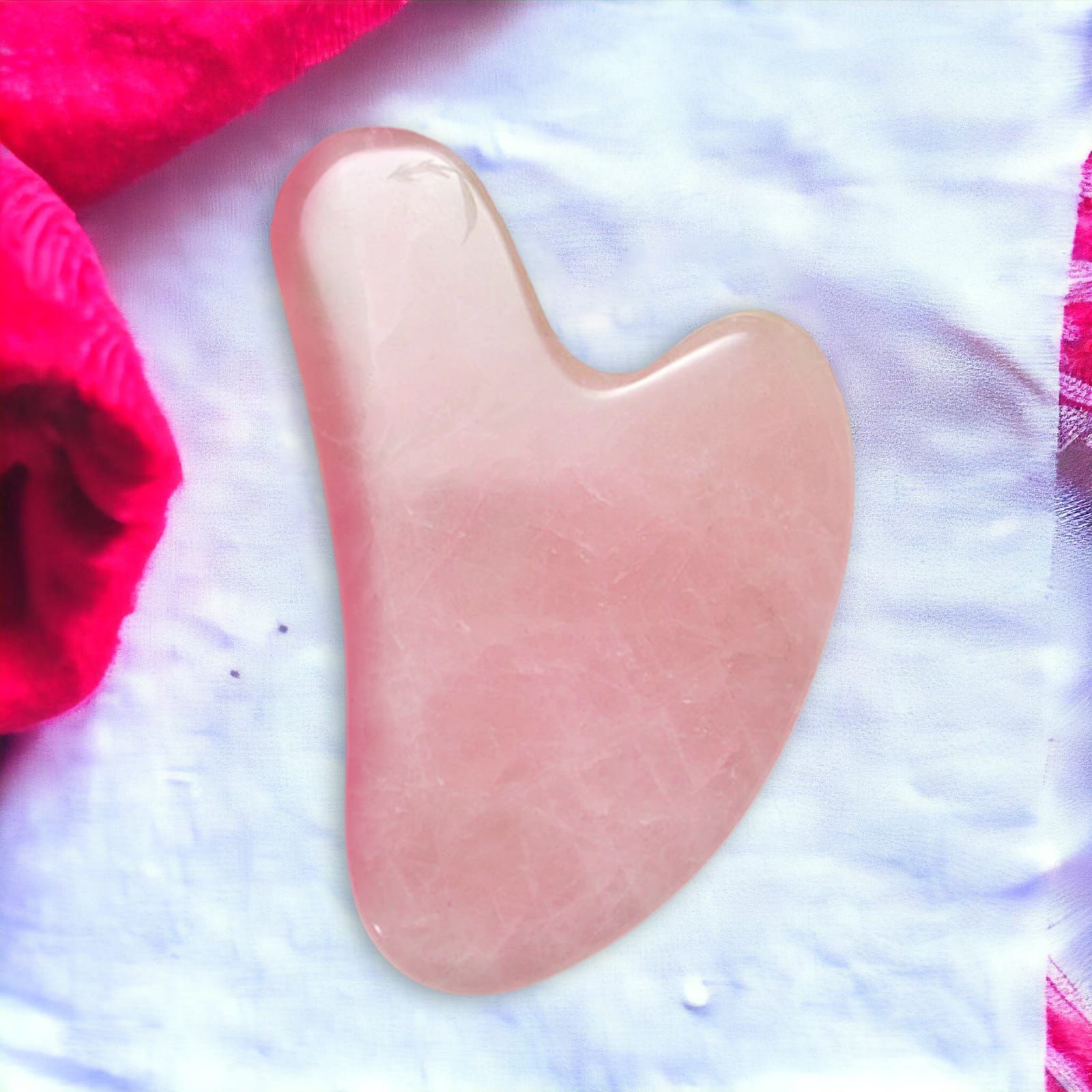 Gua Sha rosa in pietra, a forma di cuore. Su tessuto bianco, accanto a un asciugamano rosso.