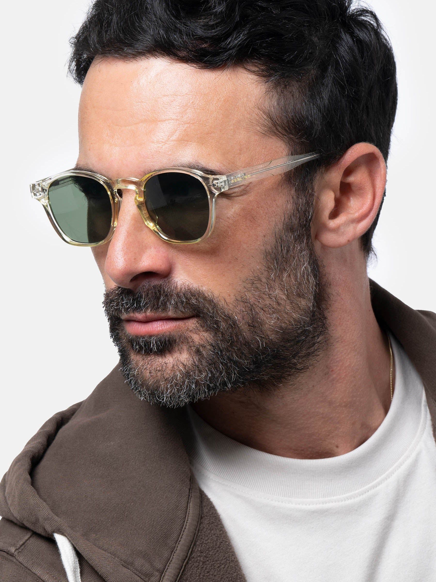 Uomo con occhiali da sole con montatura trasparente e lenti verdi. 'Herling' è visibile sull'asta.