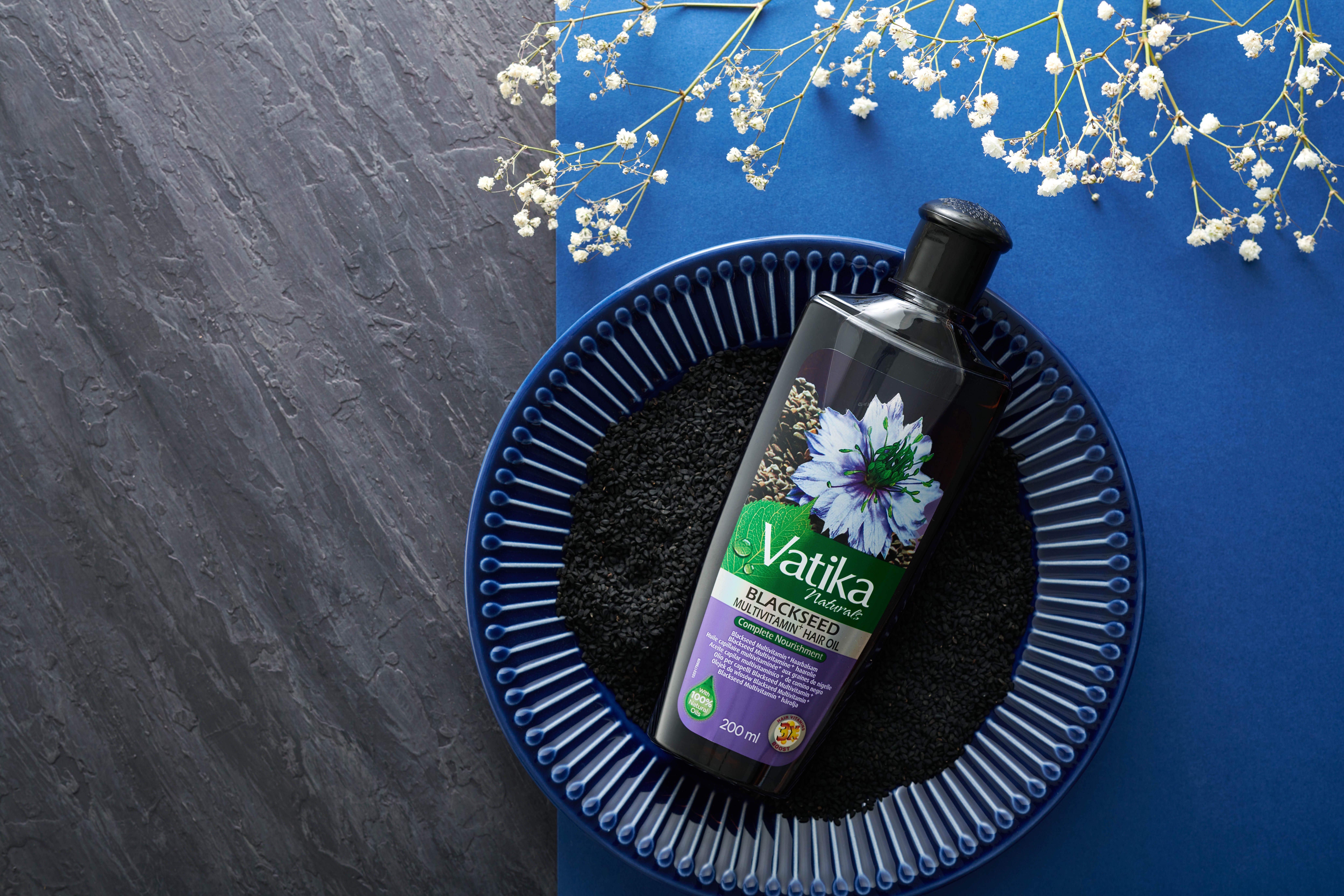 Olio per capelli Vatika Naturals Schwarzkümmel su piatto blu con semi di cumino nero. Su sfondo blu e grigio.