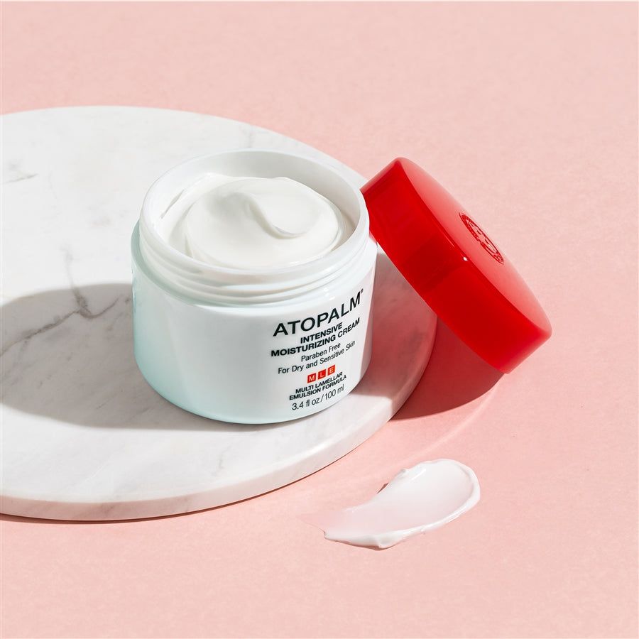 Vasetto bianco aperto con coperchio rosso. Scritta: ATOPALM INTENSIVE MOISTURIZING CREAM. Crema su lastra di marmo e sfondo rosa.