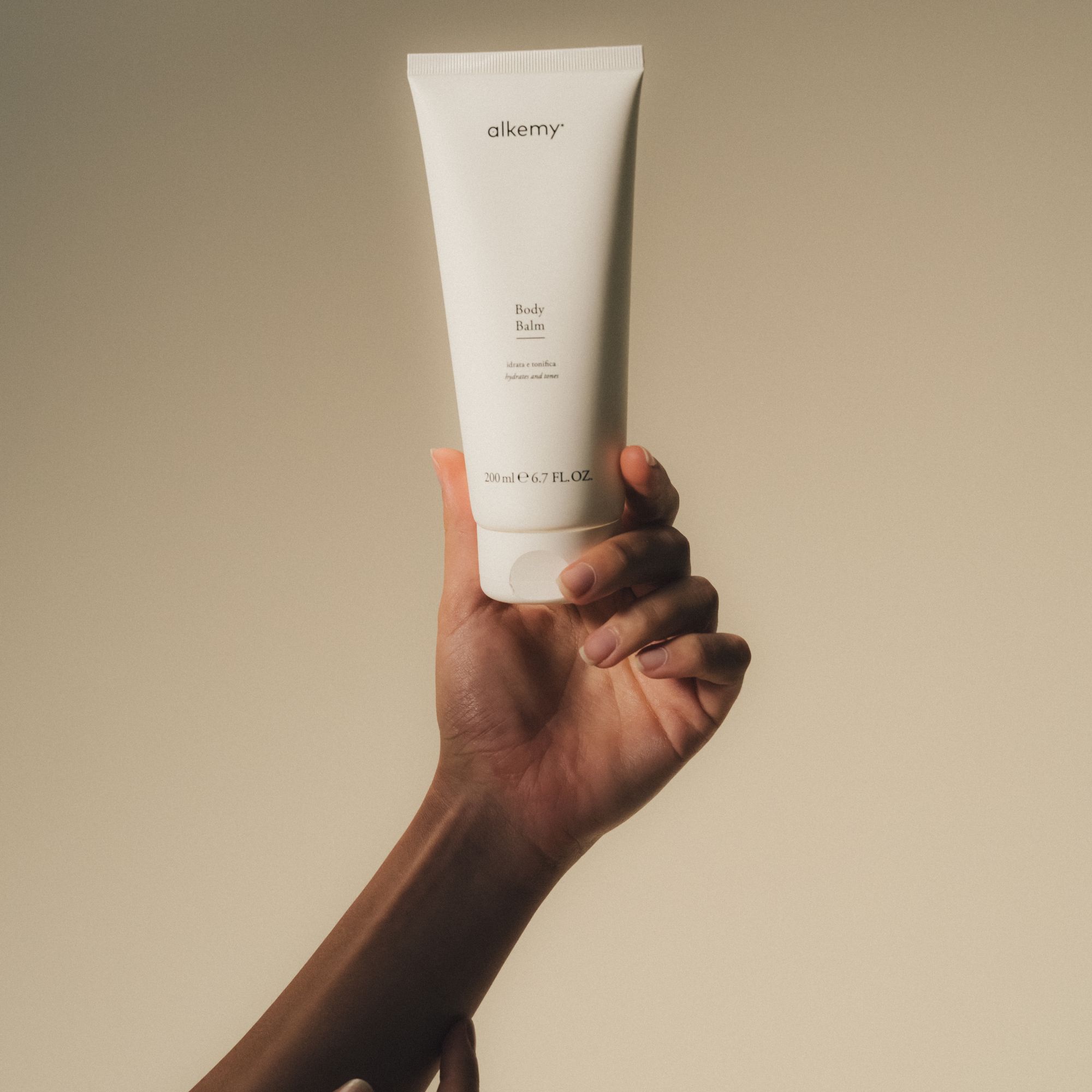 Una mano tiene un tubetto bianco di balsamo corpo. Il tubetto ha un tappo a scatto e il logo 'alkemy'.