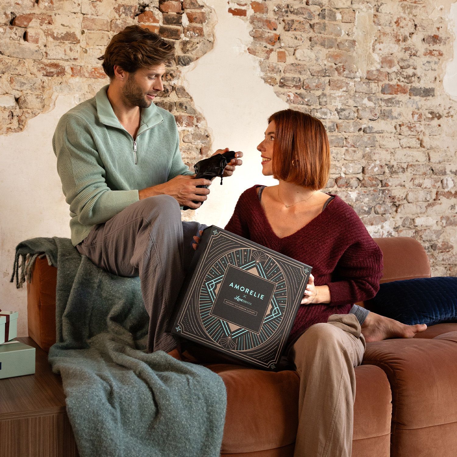 Un uomo e una donna sono seduti insieme. La donna tiene in mano una scatola del calendario dell'Avvento. Logo AMORELIE visibile.