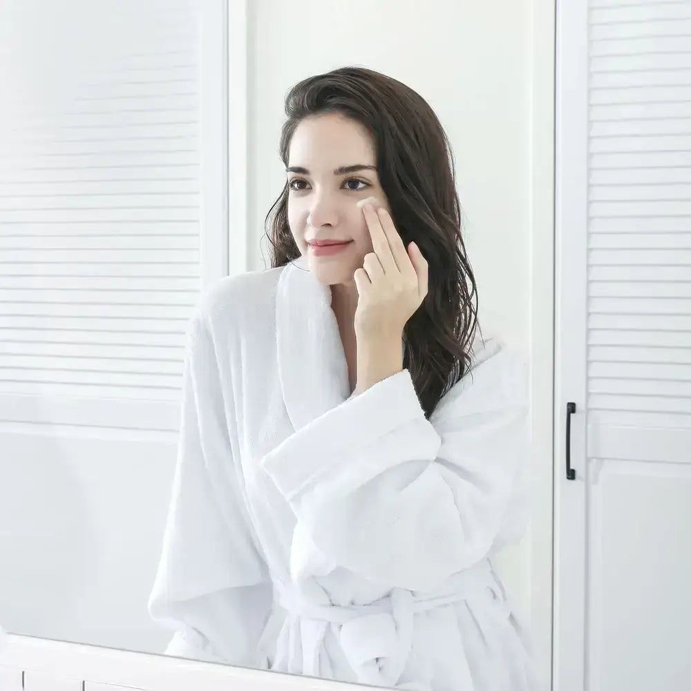 Donna in accappatoio bianco applica crema per gli occhi. Specchio sullo sfondo. Ambiente bagno luminoso.