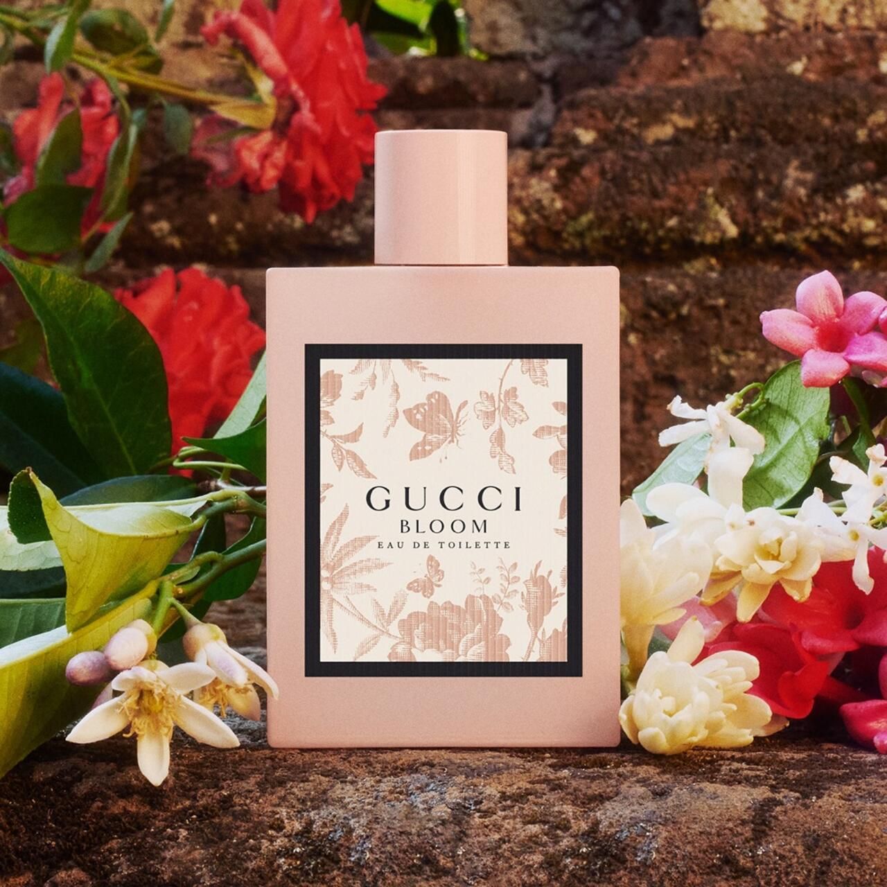 Flacone rosa con stampa floreale, davanti a un muro di pietra e fiori. Scritta: Gucci Bloom Eau de Toilette.