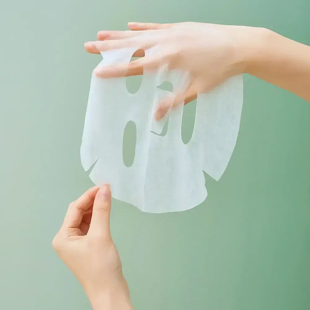 Maschera viso bianca, con fori per occhi, naso e bocca. Mani che tengono la maschera su sfondo verde.
