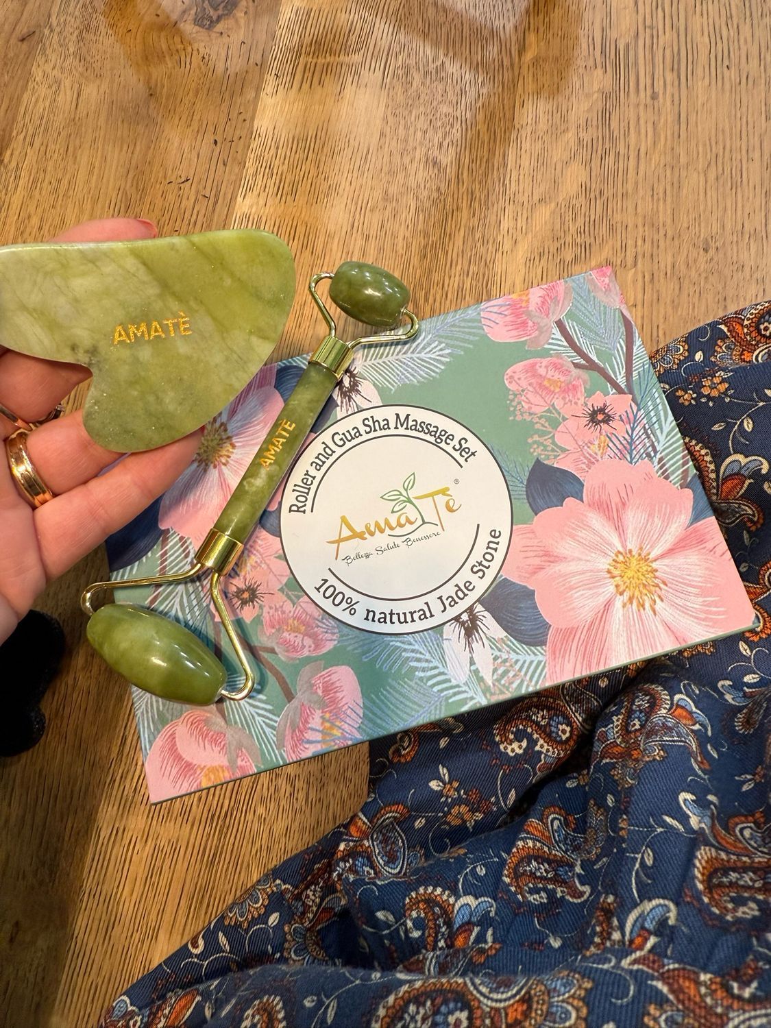 Rullo e pietra Gua Sha in giada accanto alla confezione. Confezione floreale con logo AMATÈ. Mano che tiene i prodotti.