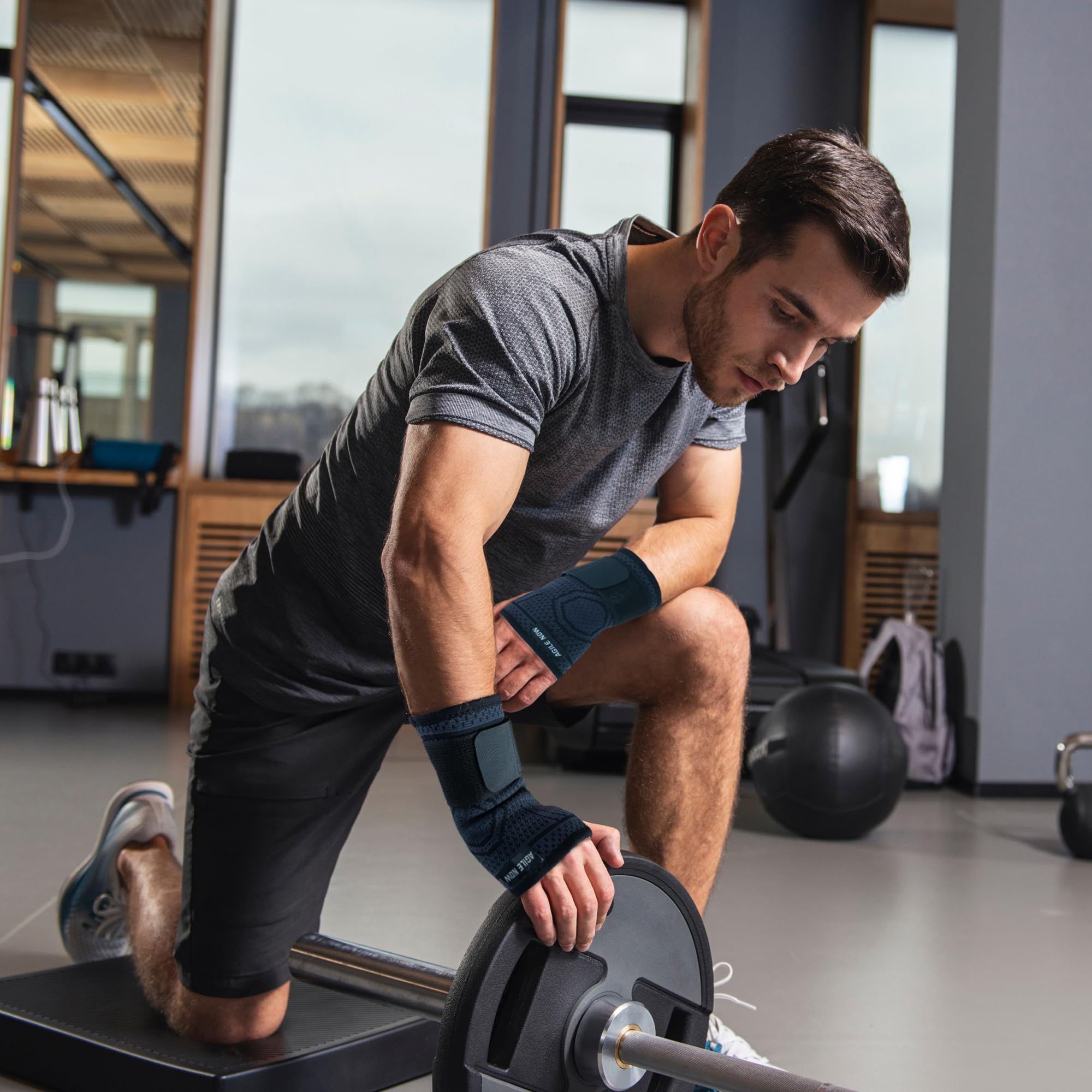 Uomo con tutore per polso durante l'allenamento. Nero. Testo: AGILE NOW. Con manubrio. Palestra sullo sfondo.