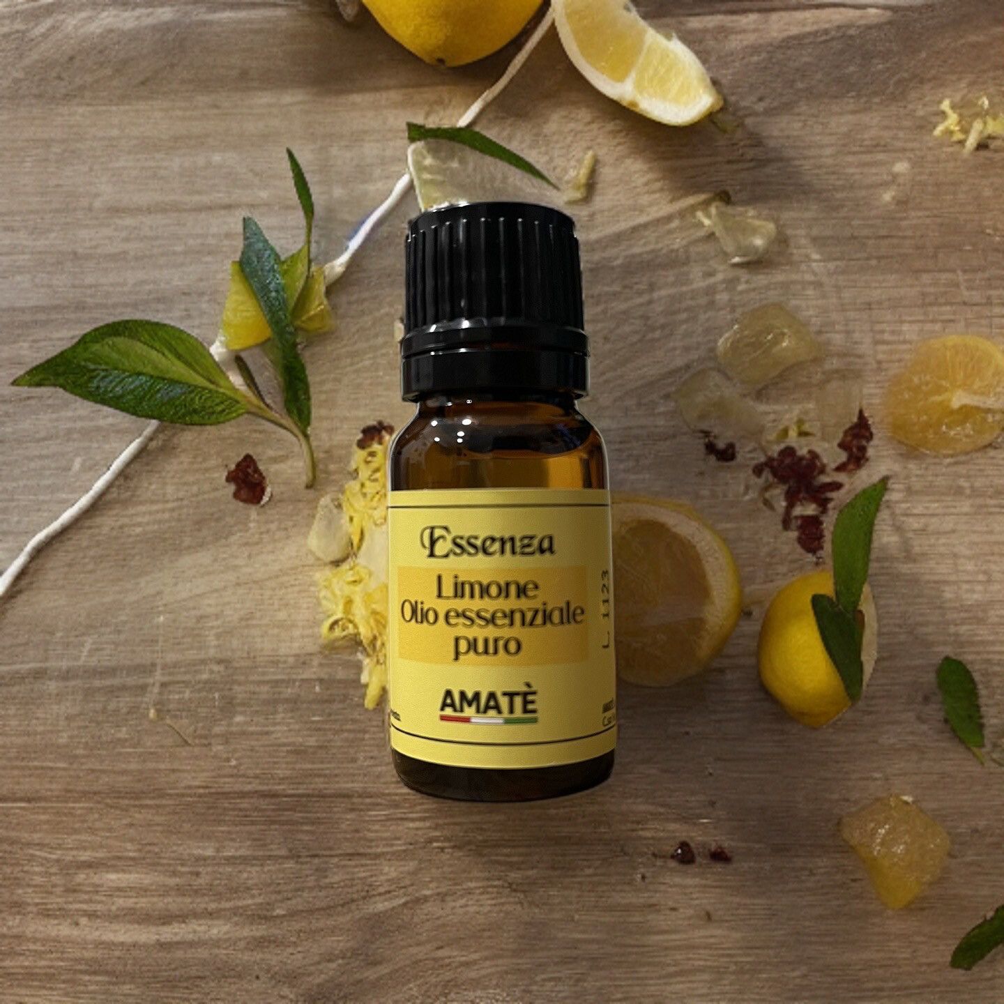Flacone di vetro marrone con tappo nero, circondato da limoni, fiori e foglie. Etichetta gialla con testo: Essenza Limone, Olio essenziale puro. Logo AMATÈ.