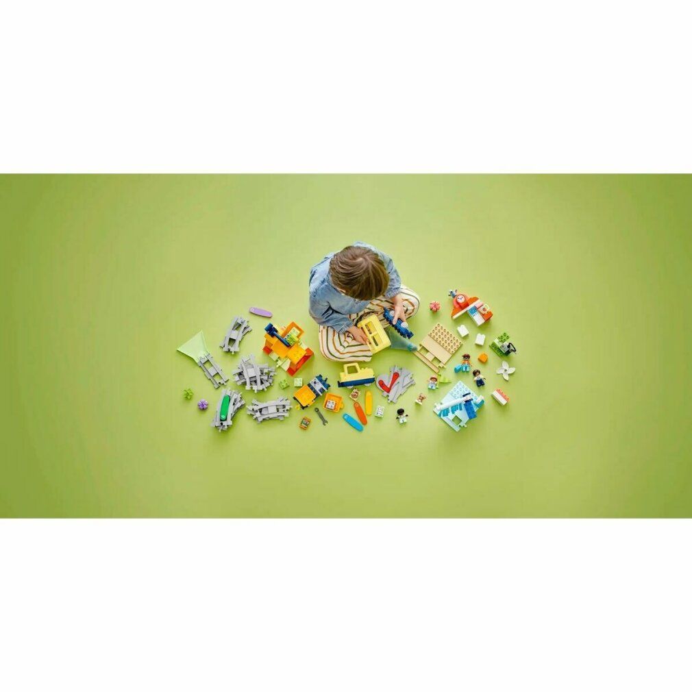 Bambino seduto su sfondo verde, circondato da pezzi LEGO Duplo. Treno giallo e accessori.