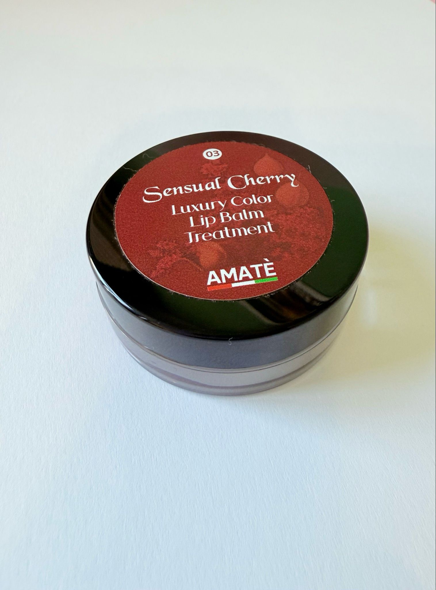 Vasetto rotondo con coperchio nero. Sul coperchio, la scritta "Sensual Cherry Luxury Color Lip Balm Treatment" e "AMATÈ".