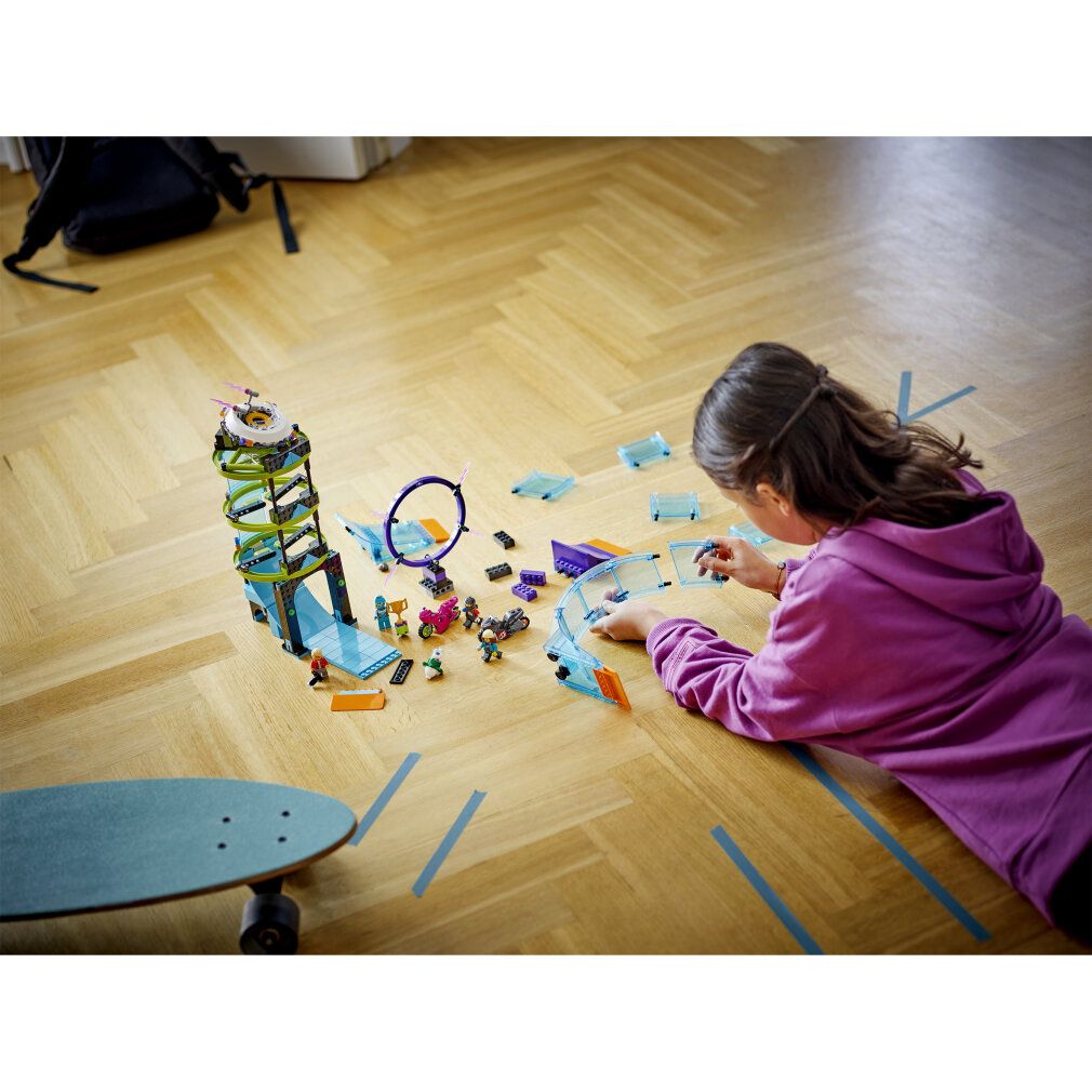 Bambina che costruisce il set LEGO City Stuntz su pavimento in legno. Pezzi e personaggi sparsi. Skateboard in primo piano.