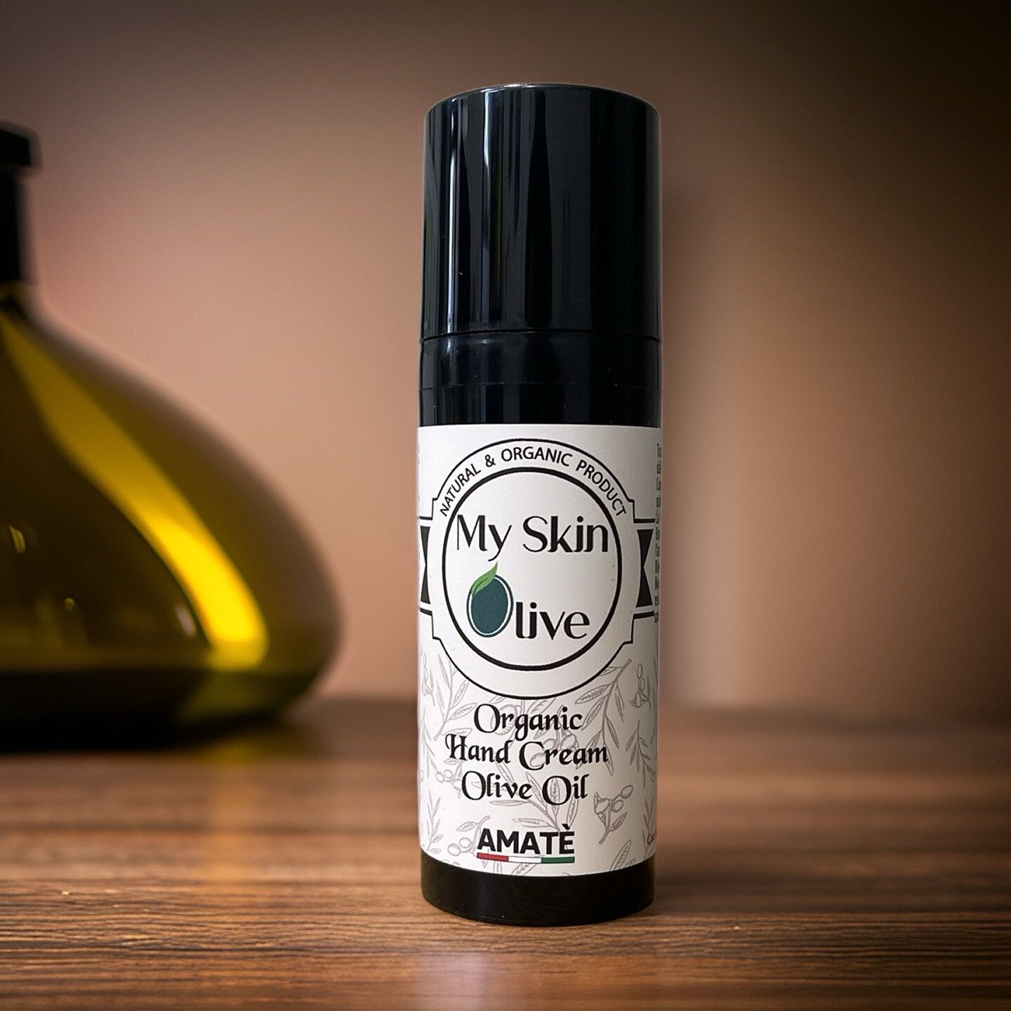 Flacone nero su sfondo sfocato. Scritta: My Skin Olive, Organic Hand Cream Olive Oil, Amatè.