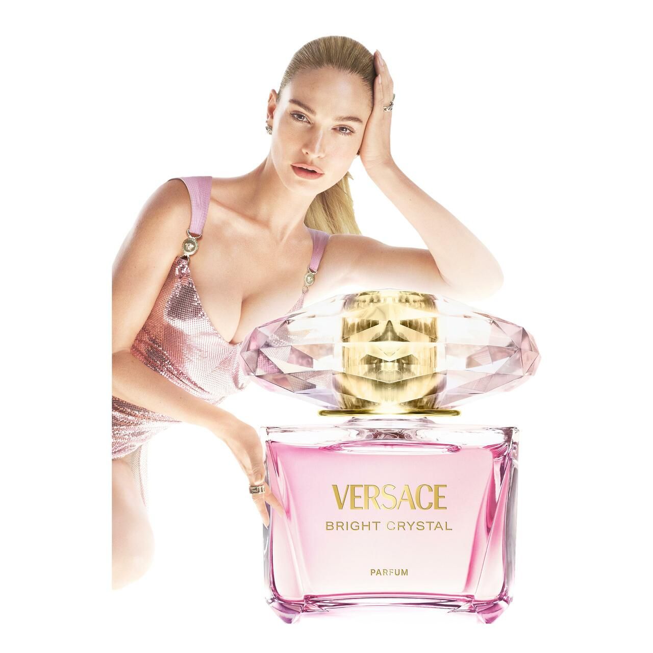 Flacone di profumo rosa. Donna appoggiata al flacone. Contenitore rettangolare con tappo dorato e cappuccio di cristallo. Scritta Versace.
