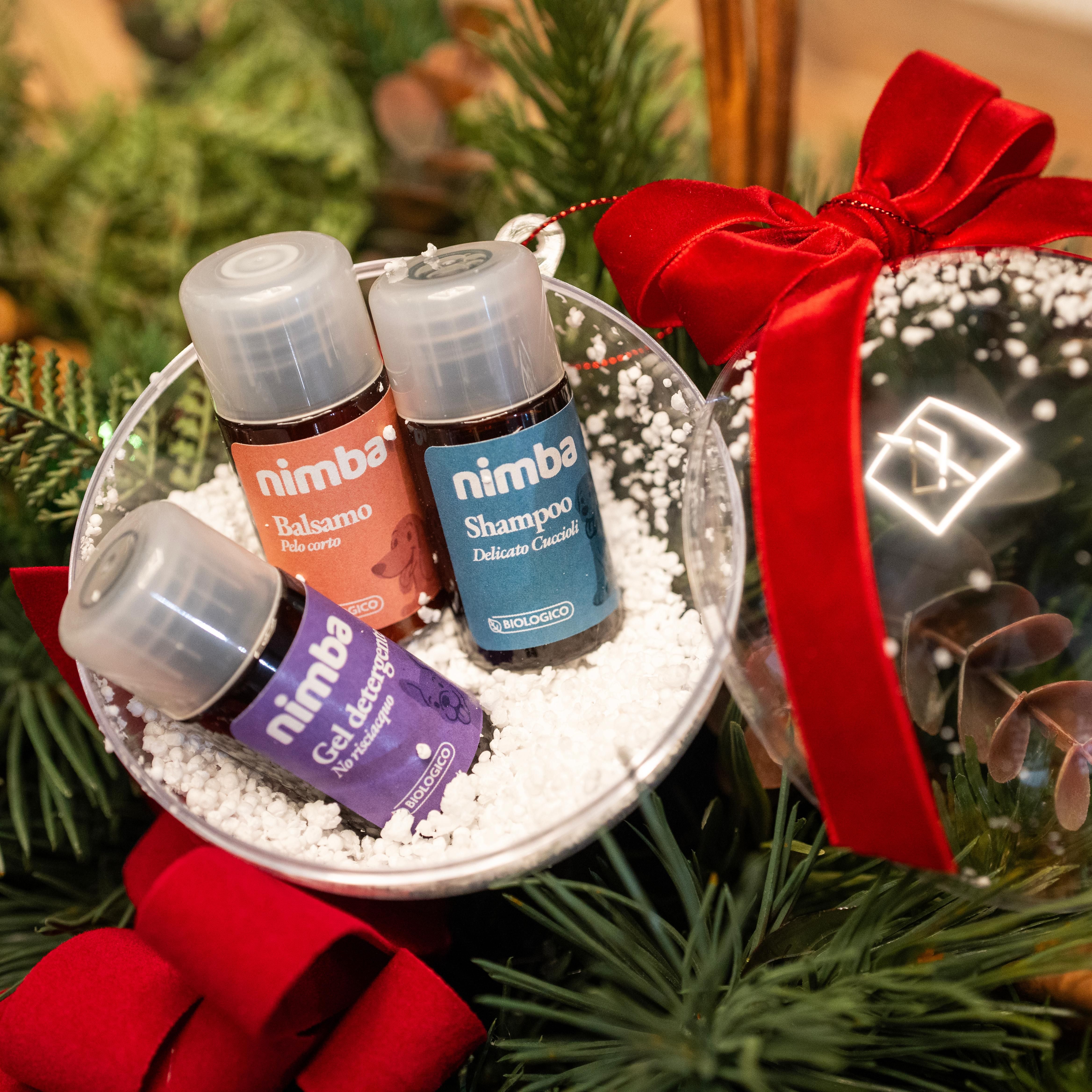 Ornamento natalizio con tre flaconi "nimba" nella neve finta. Fiocco rosso e sfondo di rami di pino.
