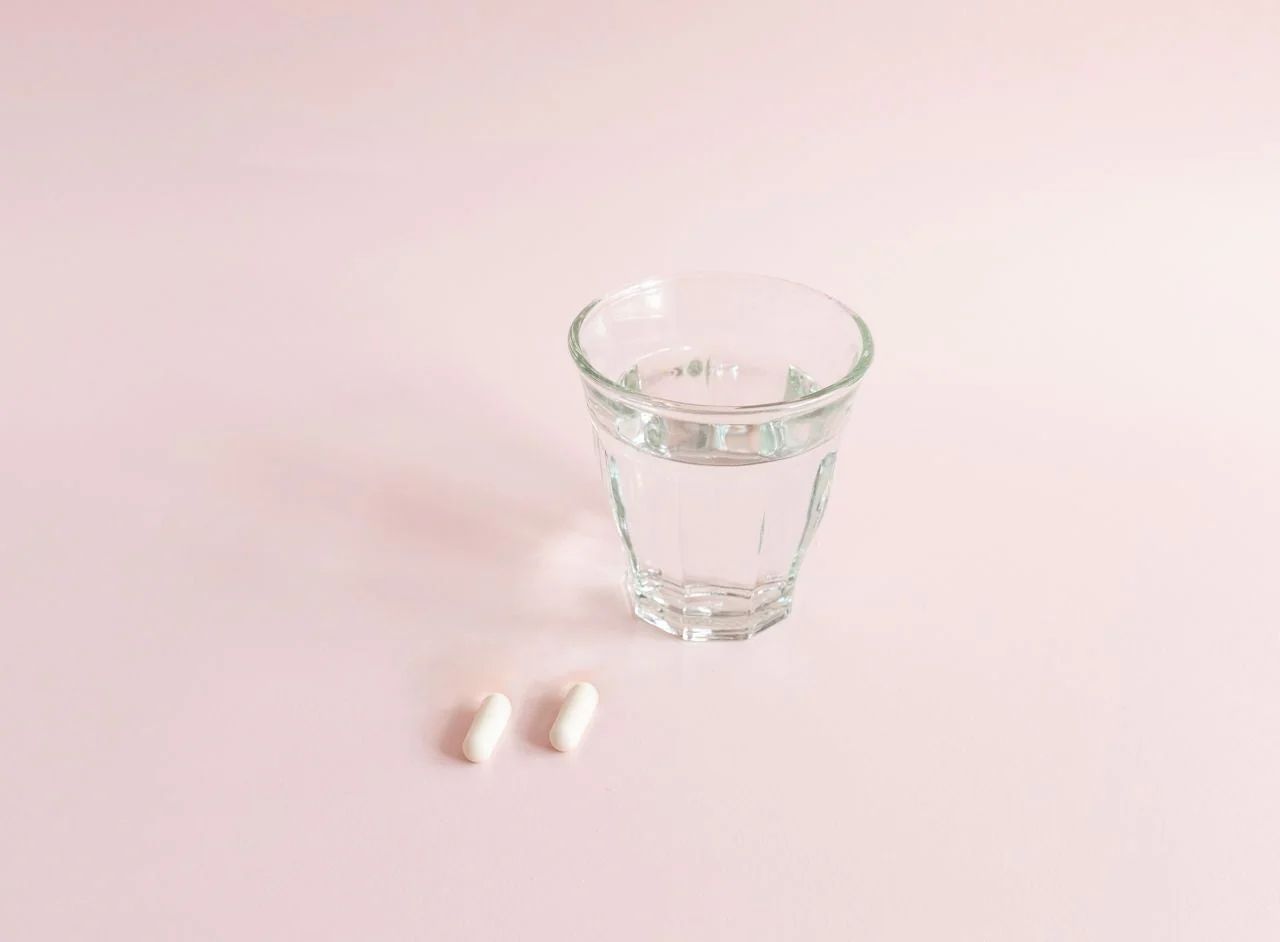 Bicchiere d'acqua e due capsule bianche su sfondo rosa.