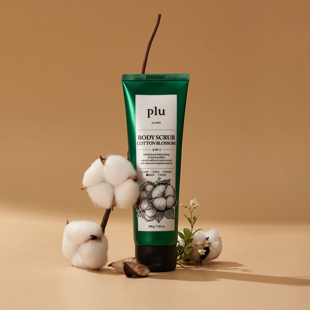 Tubo verde con etichetta bianca. Scritta: PLU Body Scrub Cotton Blossom. Ramo di cotone e fiori accanto al tubo.