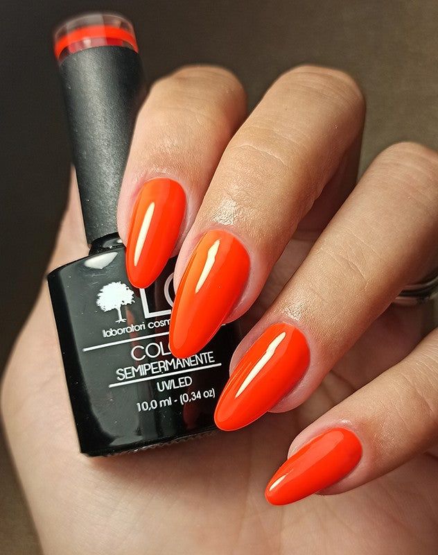 Mano con unghie smaltate e flacone di smalto. Unghie smaltate arancione neon. Flacone con tappo nero. Testo: LCI, Colore Semipermanente.