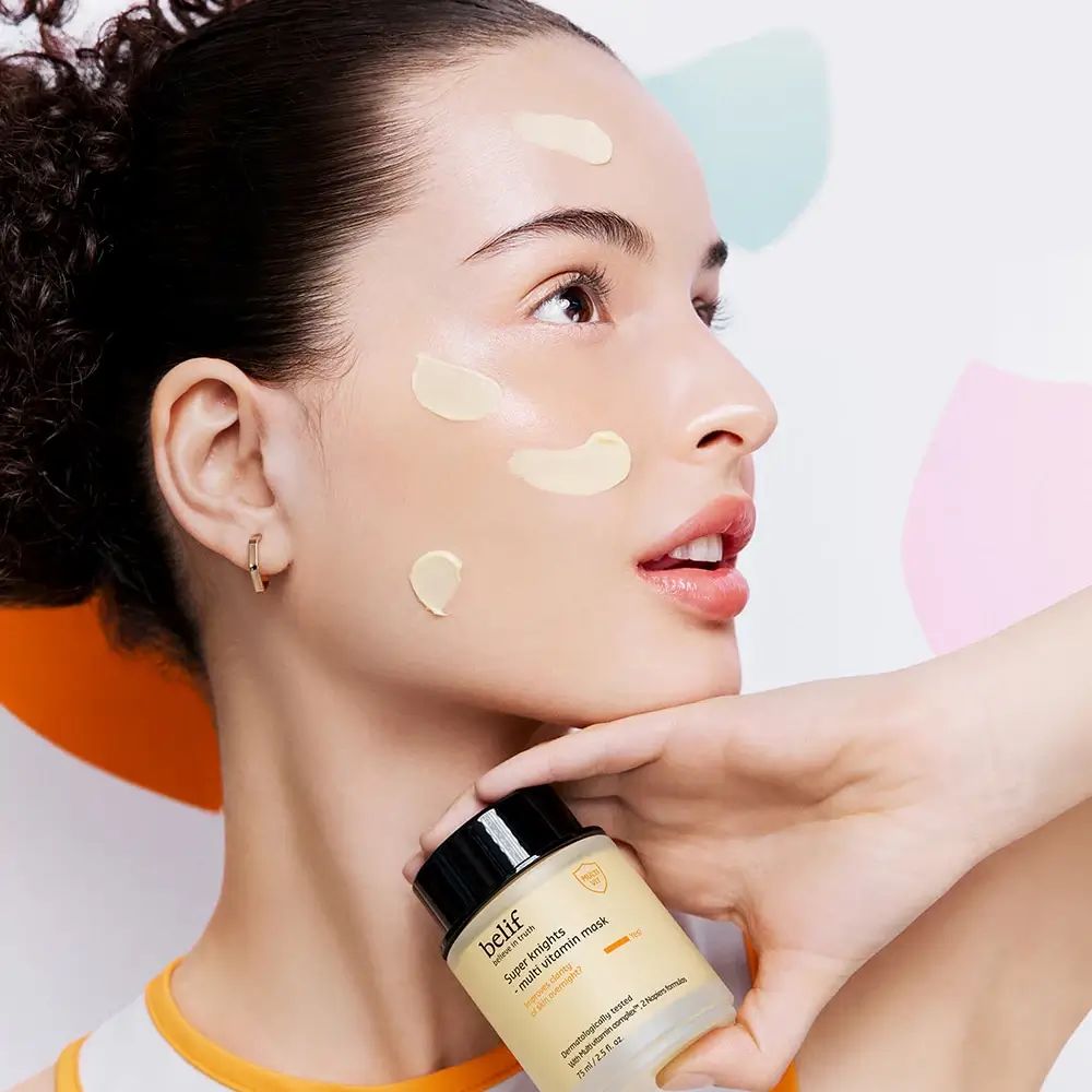 Donna con maschera sul viso, tiene il prodotto. Belif Super Knights Multi-Vitamin Mask. Crema giallastra sulla pelle. Sfondo con forme colorate.